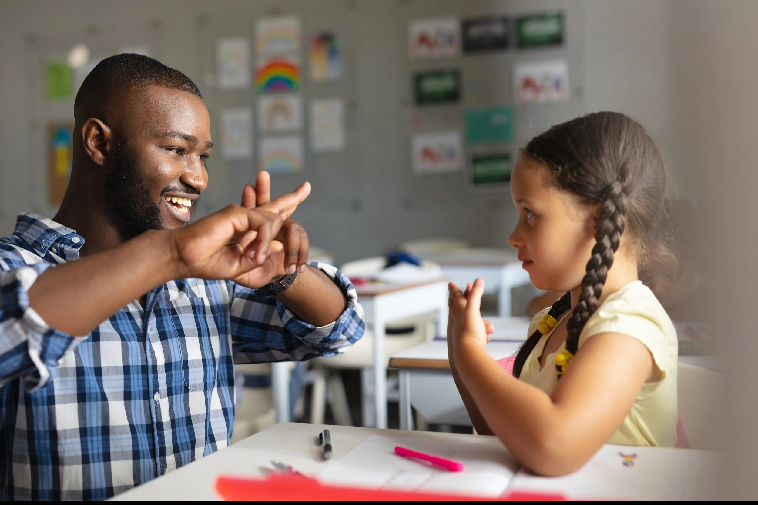 A man and a young girl sitting at a desk in a classroom, engaging in a playful finger gesture activity. The man is smiling and wearing a blue plaid shirt, while the girl is focused, with braided hair and a yellow shirt. The classroom background is de