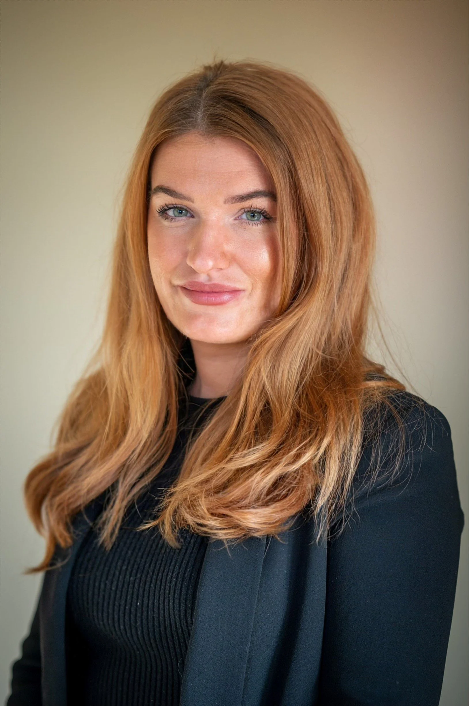 A woman with long, wavy red hair and blue eyes wearing a black top and black blazer.