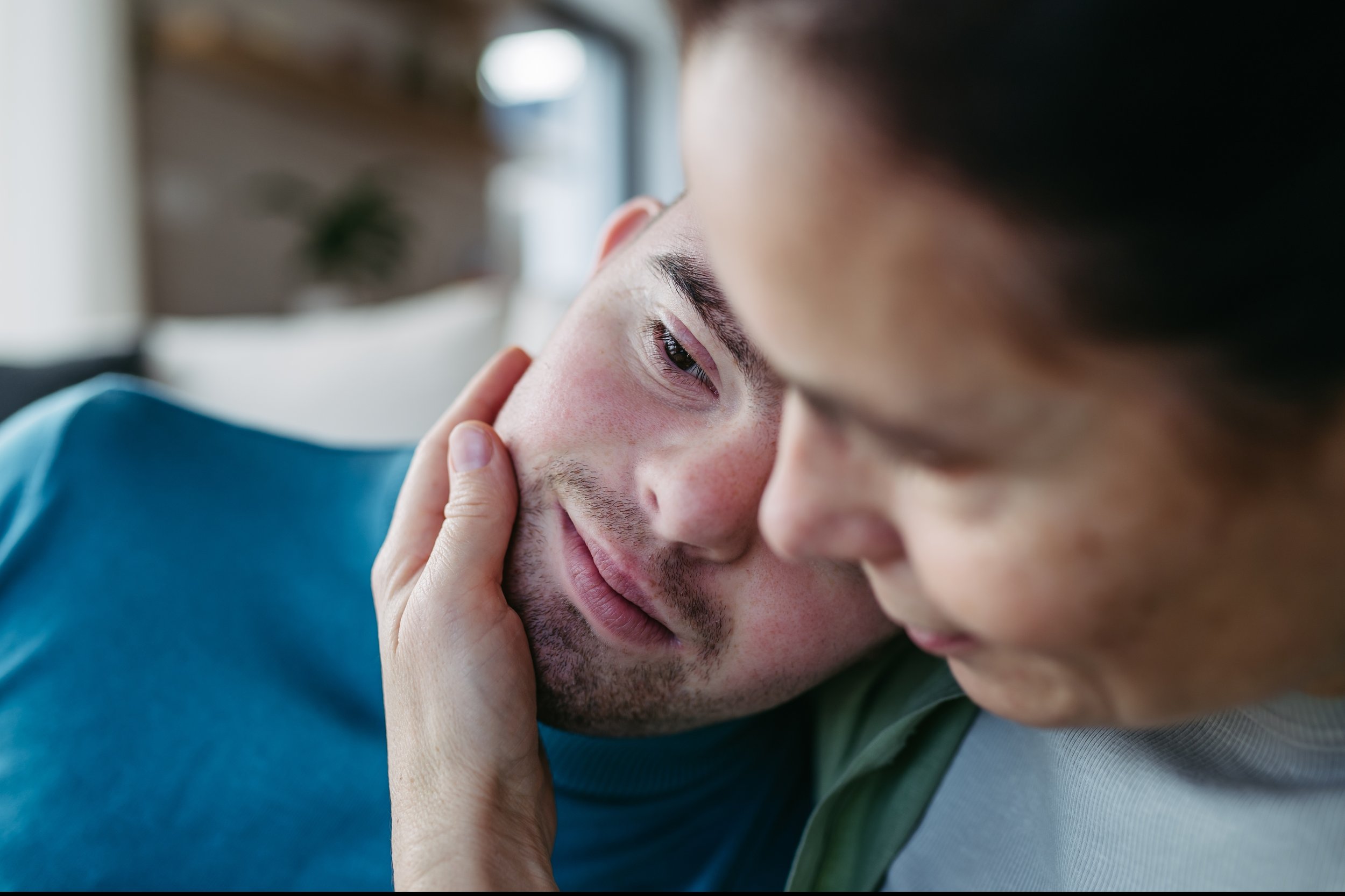 Close-up of two men, one gently holding the other's face, sharing an intimate moment indoors.