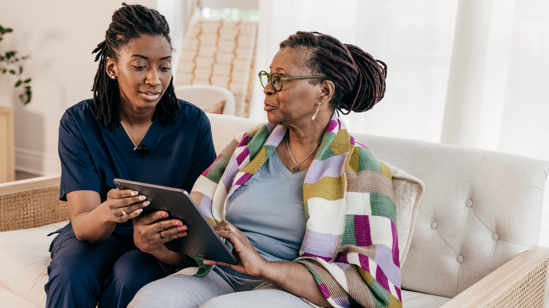 A caregiver and an elderly woman look at a tablet together on a sofa in a bright living room.