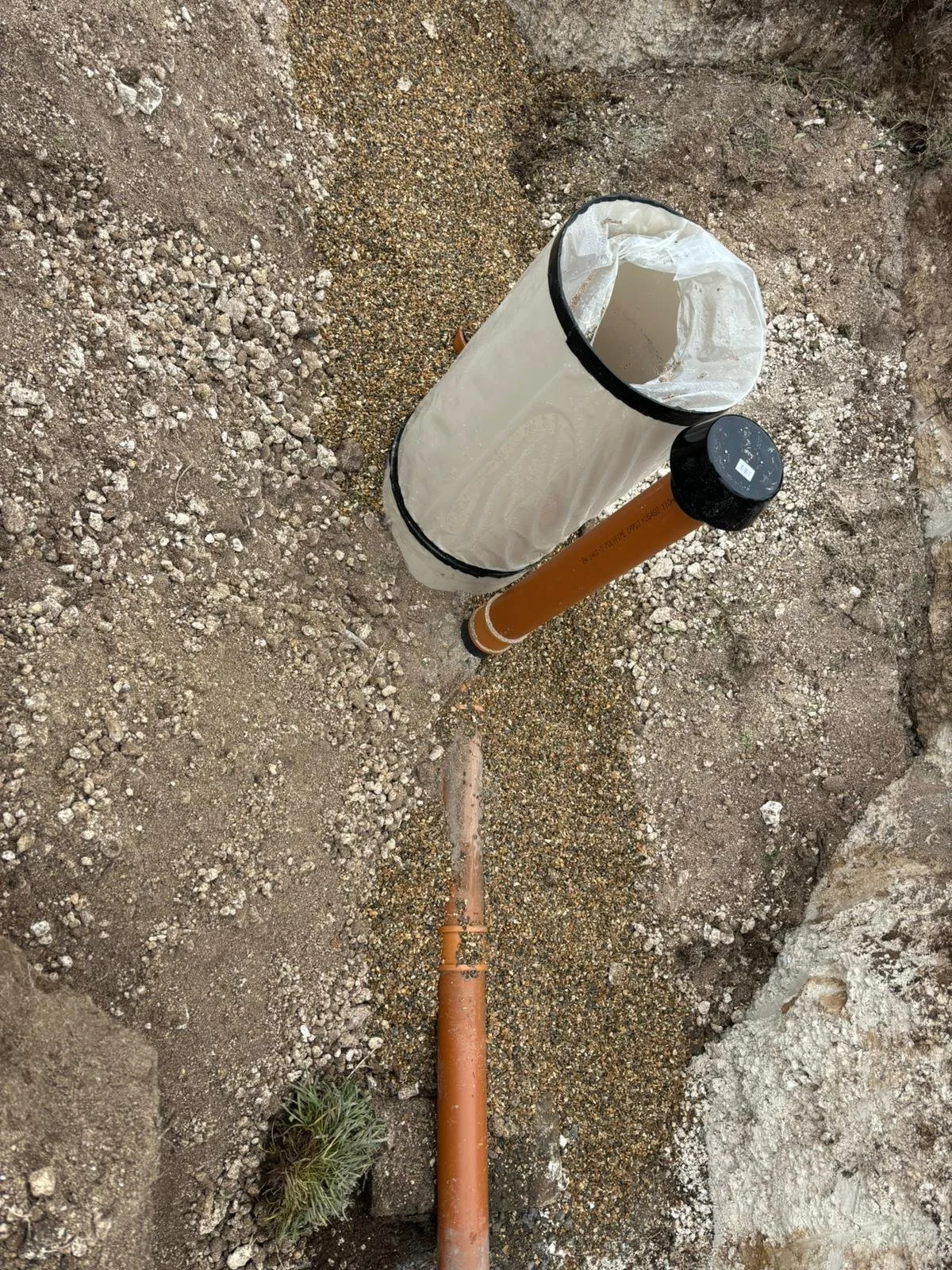 A construction cone with a bag over it, placed on a dirt ground next to an orange drainage pipe and gravel patches.