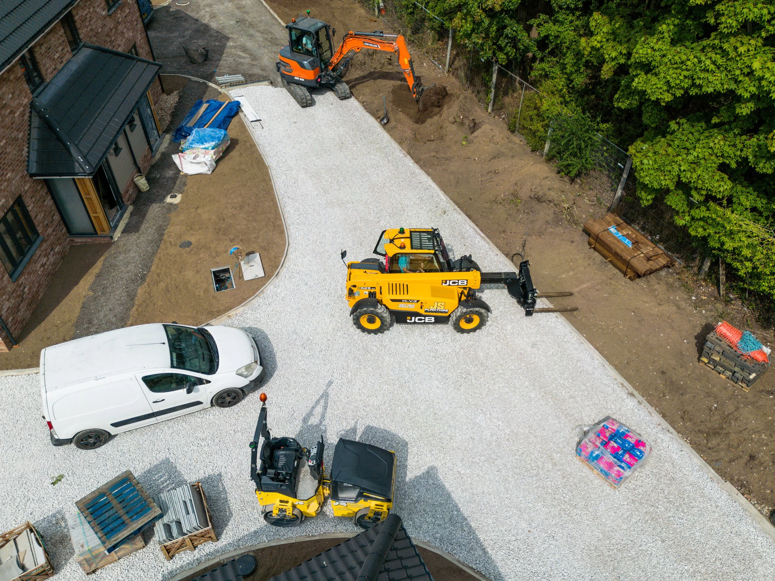 Construction site with gravel driveway, small excavator, front loader, parked car, wooden pallets, and construction materials near a brick building surrounded by green trees.