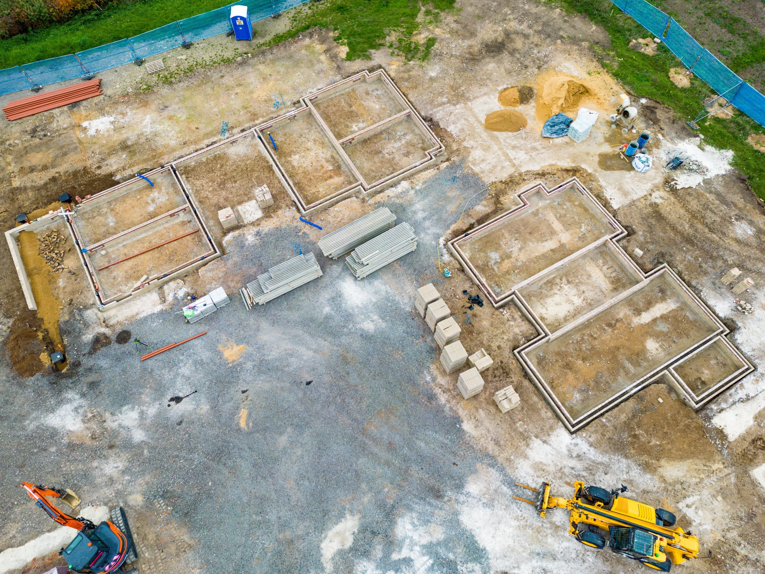 Aerial view of a construction site with partially built house foundations, construction materials, equipment, and an excavator.