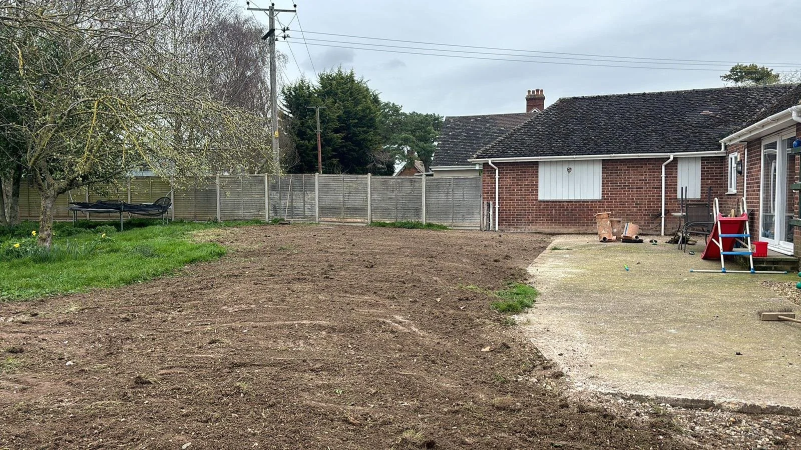 Backyard with a dirt and grass area, a tree on the left, a trampoline, a brown fence, a brick house with window blinds, and a concrete patio with children's toys and outdoor furniture.