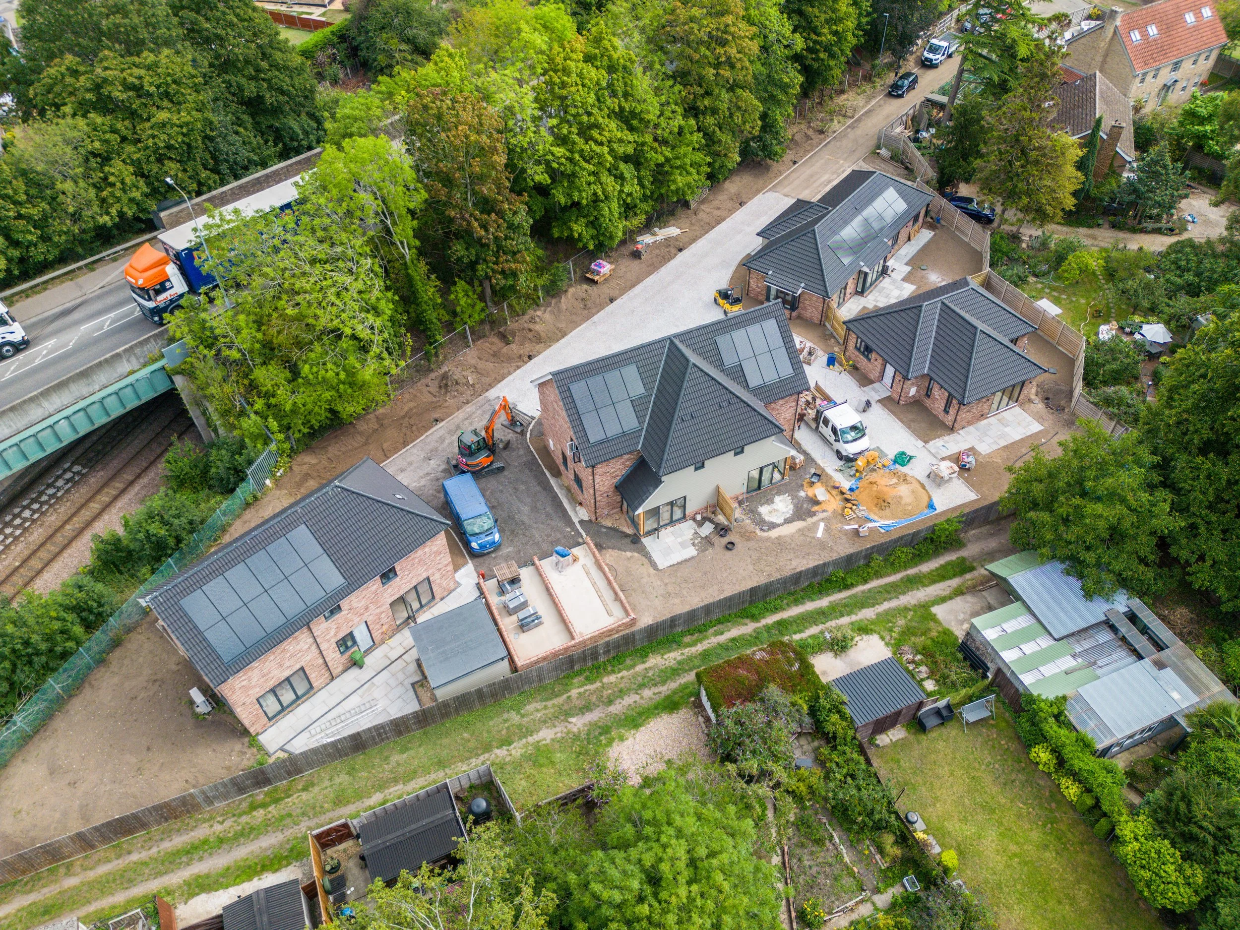 Aerial view of a construction site with several houses being built, surrounded by trees and a residential neighborhood, with construction vehicles and materials visible.
