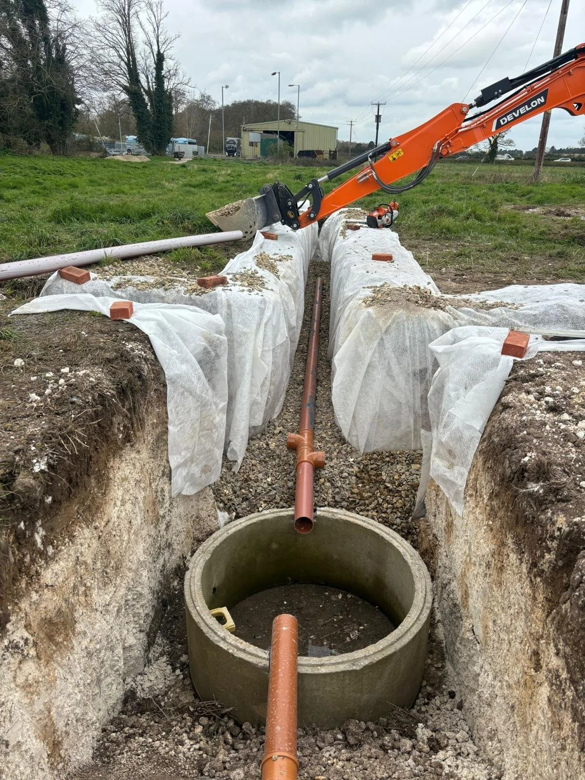 Construction site with underground pipe installation, showing a trench with a concrete manhole, orange pipes, and an excavator overhead.