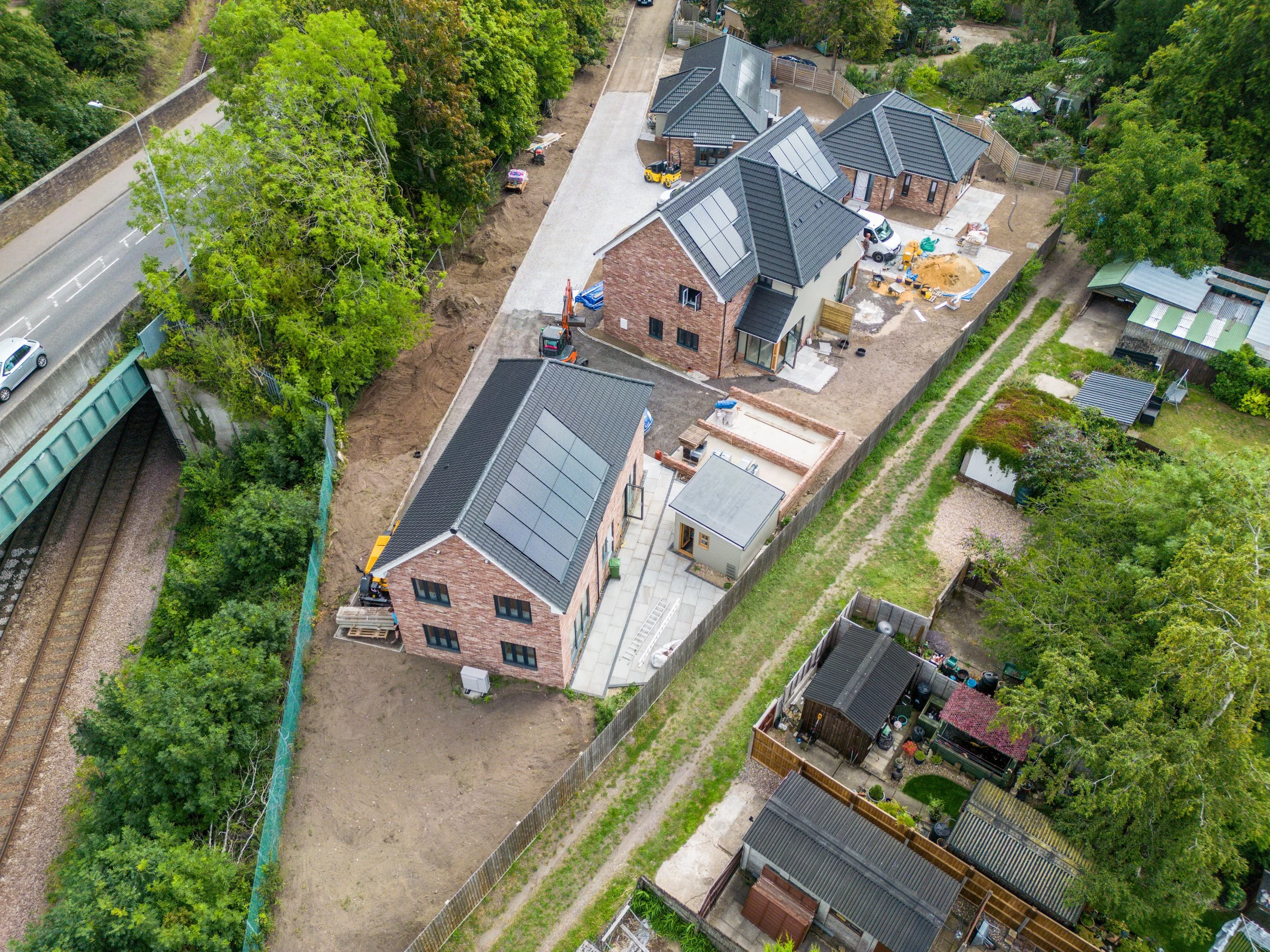 Aerial view of a row of houses under construction with solar panels on roofs, surrounded by trees, a railway, and neighboring yards.