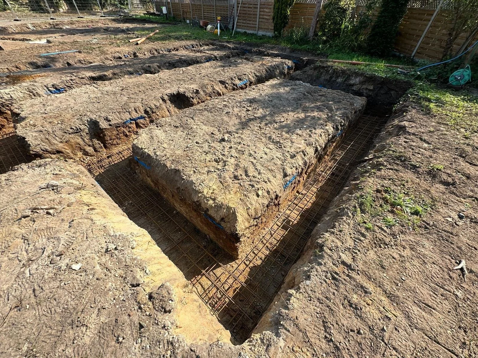 Construction site with dug-out trenches and large stone slabs, with steel rebar grid laid out in the trenches, and a wooden fence in the background.