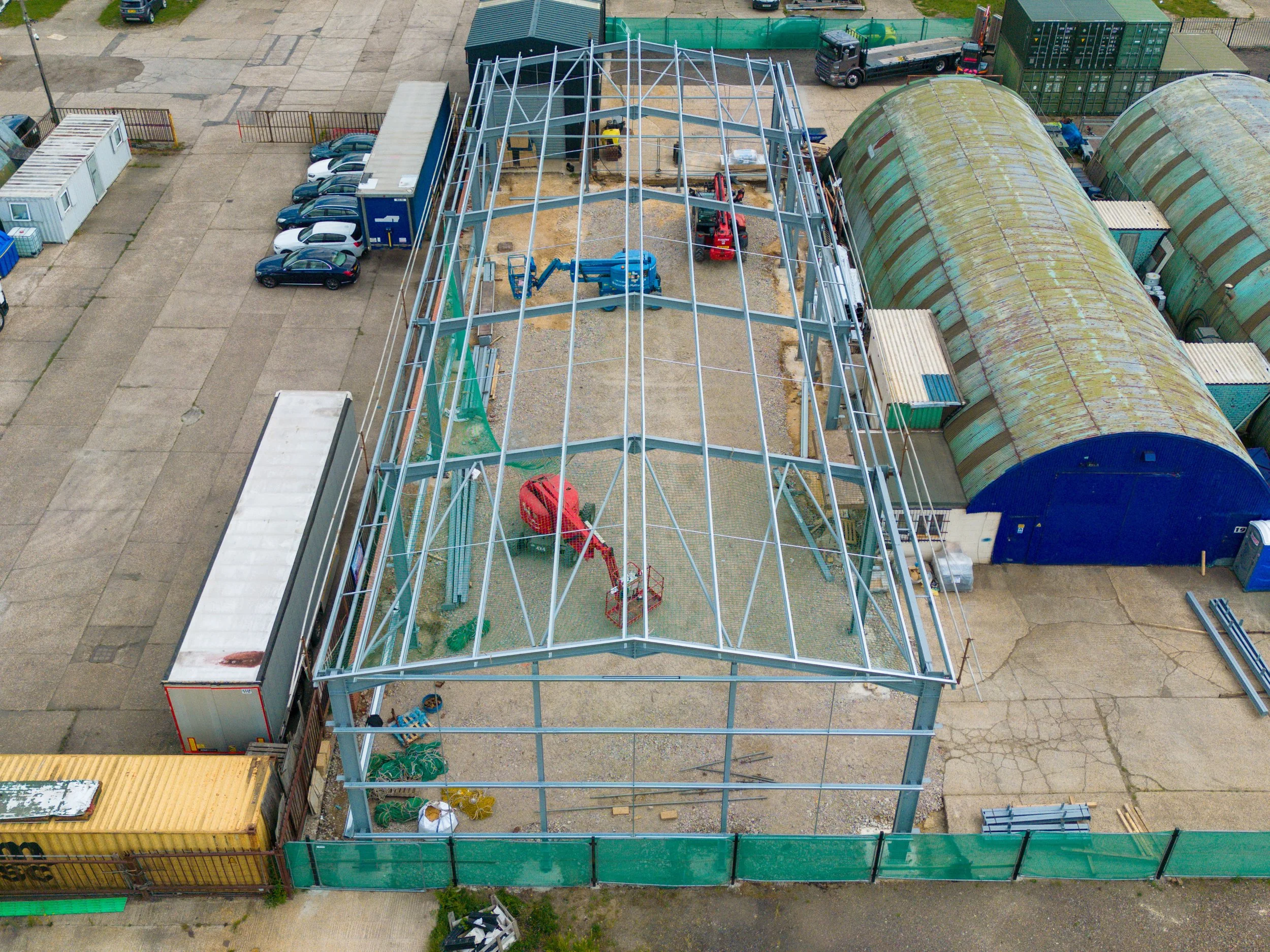 Aerial view of a construction site with a steel frame structure, adjacent to storage containers and parked cars. Two large, curved, rusted metal sheds are also visible.