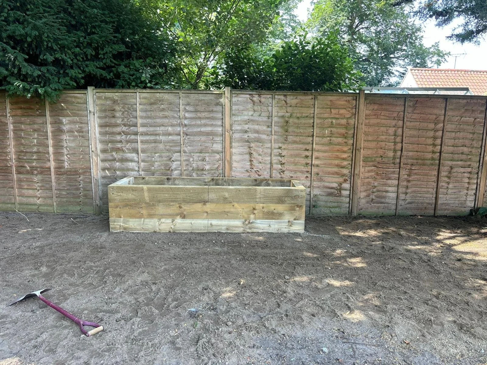 A wooden planter box in a backyard with a dirt ground and a wooden fence in the background, with some greenery above the fence and a small hammer lying on the ground.