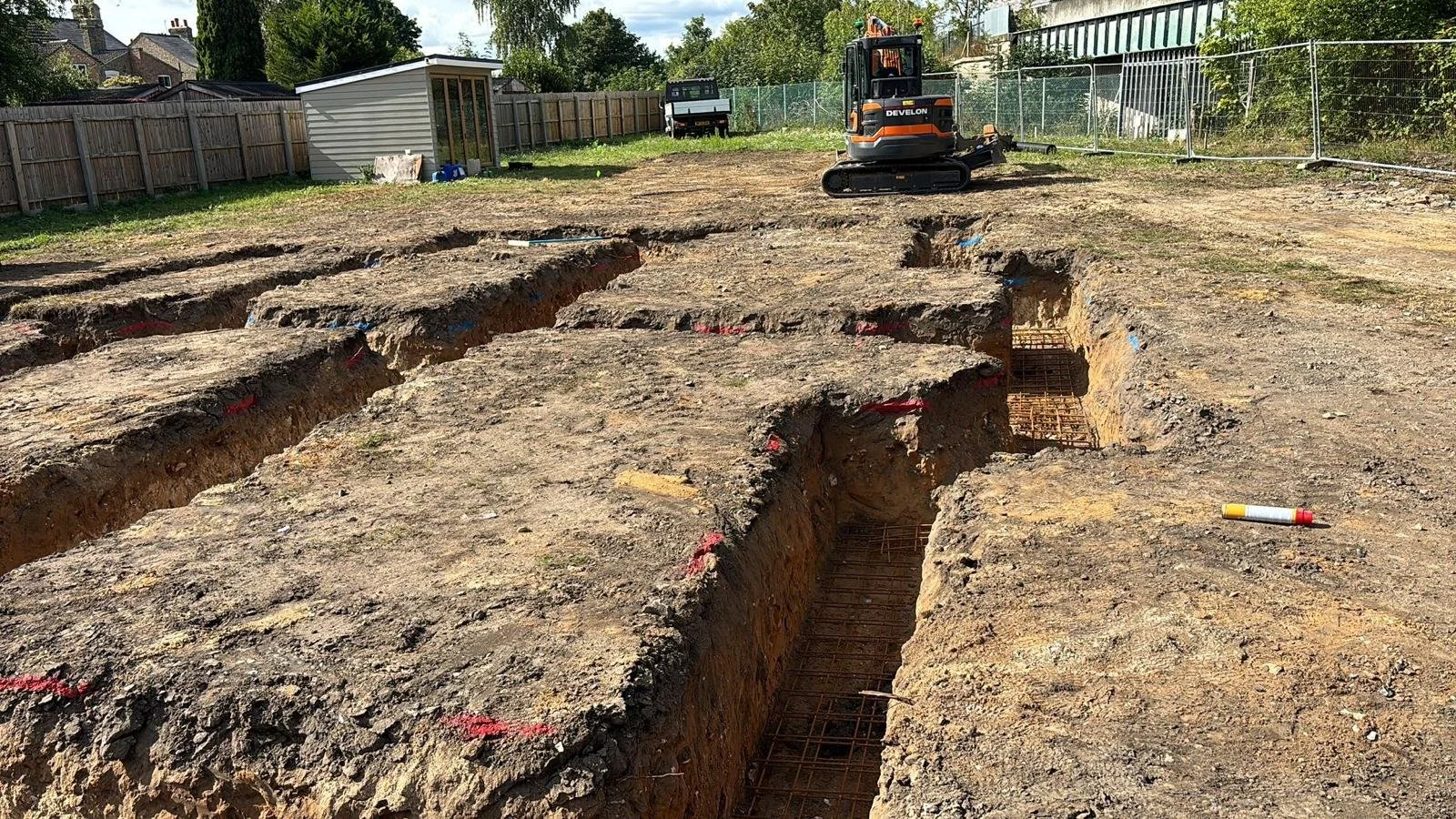 Construction site with excavated trenches and exposed rebar, a small orange tracked excavator in the background, and a fenced-off area with trees and buildings beyond.