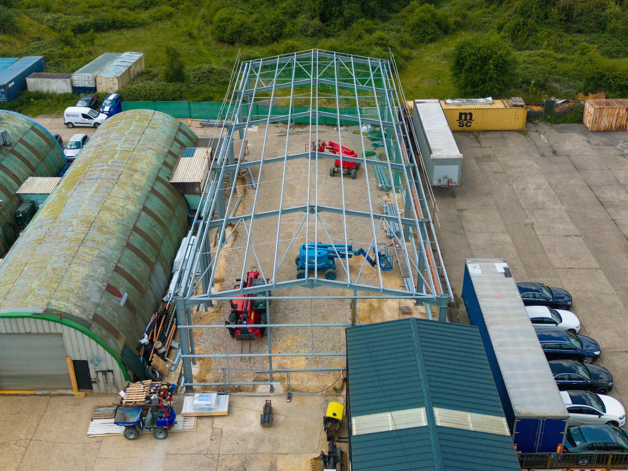Construction site with steel framework for a building, construction vehicles, and parked cars, with storage containers and a green-roofed building nearby.