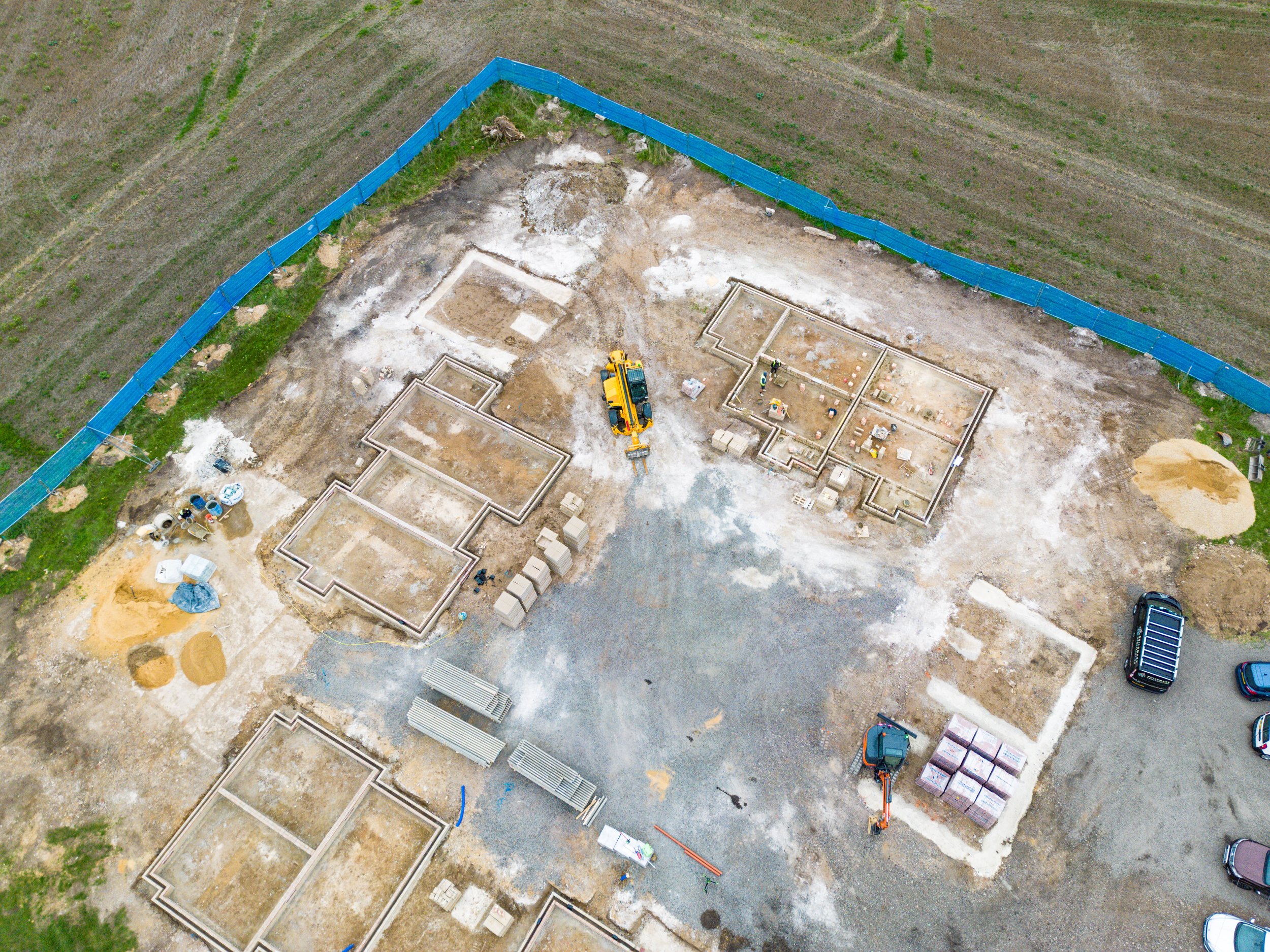 Aerial view of a construction site with foundation outlines, construction equipment, and parked vehicles, surrounded by a blue safety barrier.
