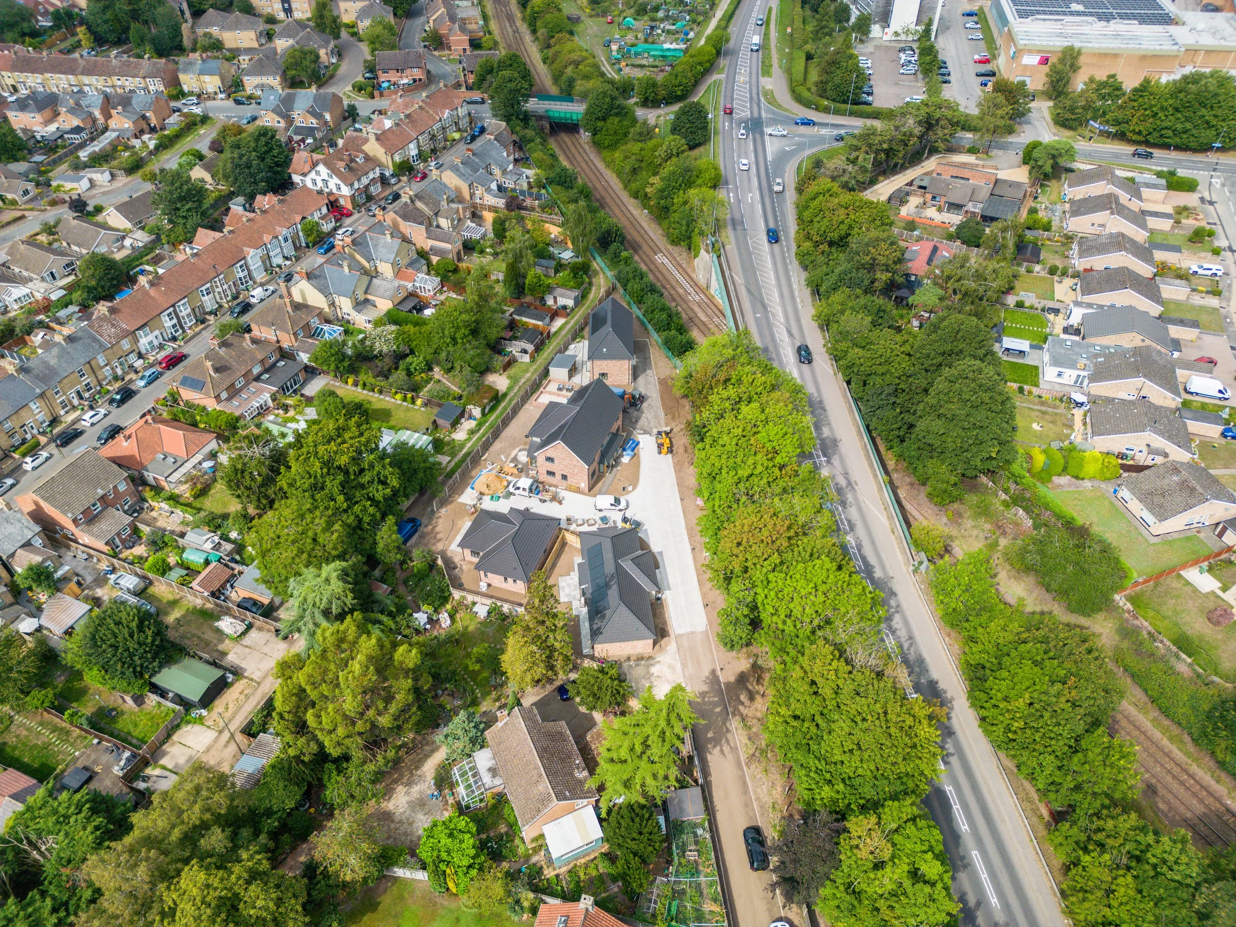 An aerial view of a suburban neighborhood with closely packed houses, trees, and streets. A railway runs through the center, with forested areas on either side and cars on the roads.