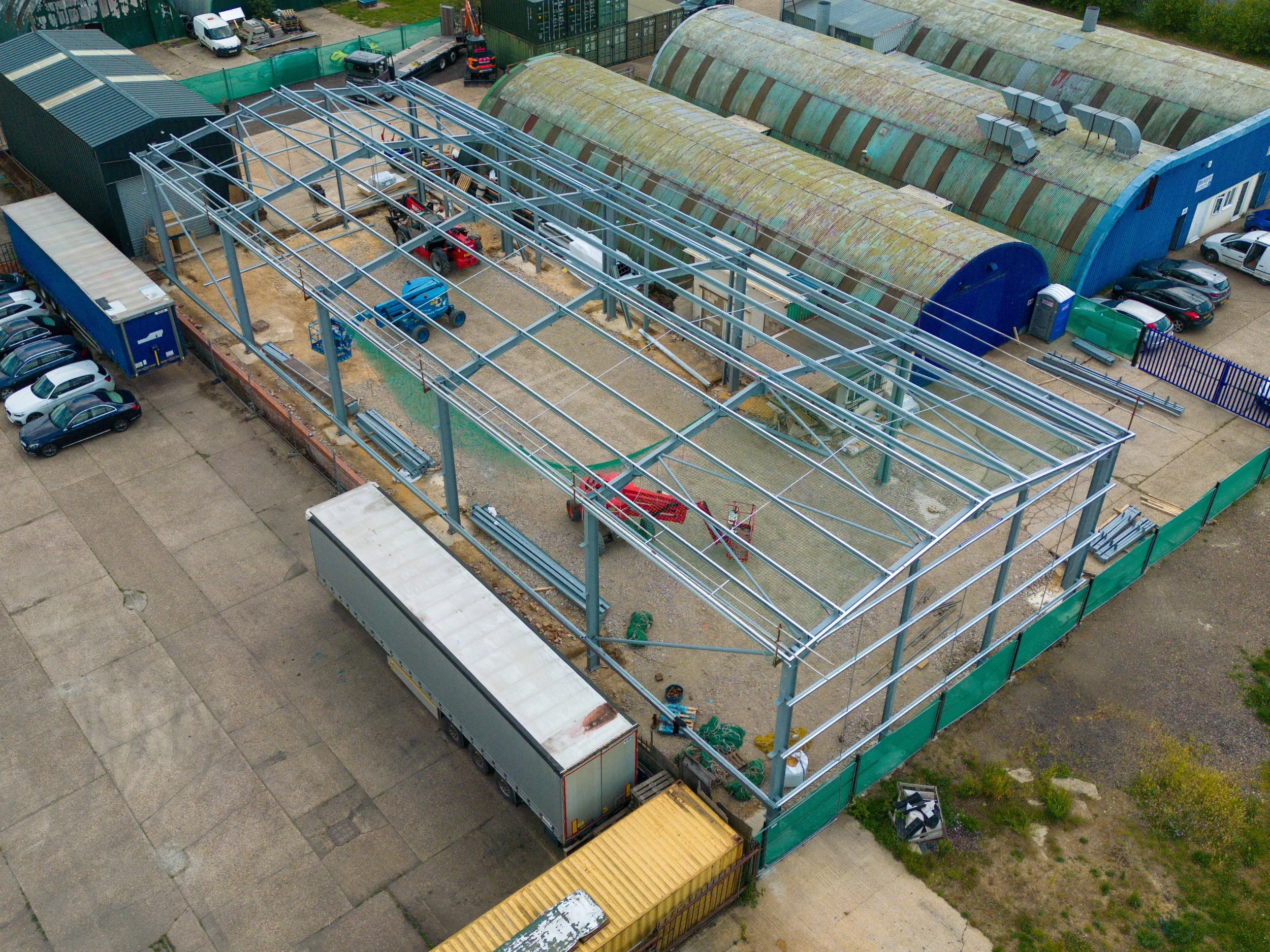 An aerial view of a construction site showing a steel framework being assembled for a building, with construction vehicles and equipment nearby, surrounded by other industrial structures and parked cars.