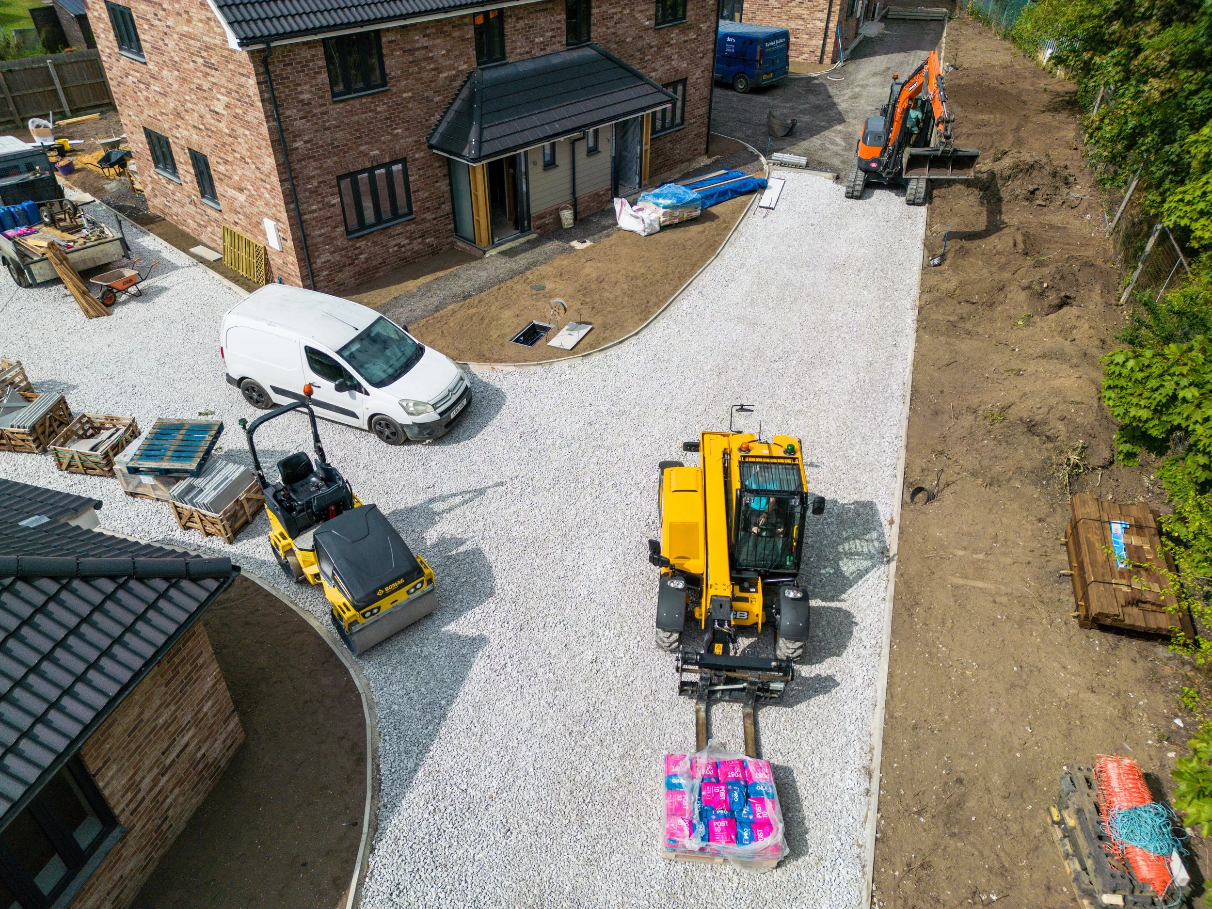 Construction site showing a new gravel driveway being laid in front of a brick house, with construction equipment and materials around.