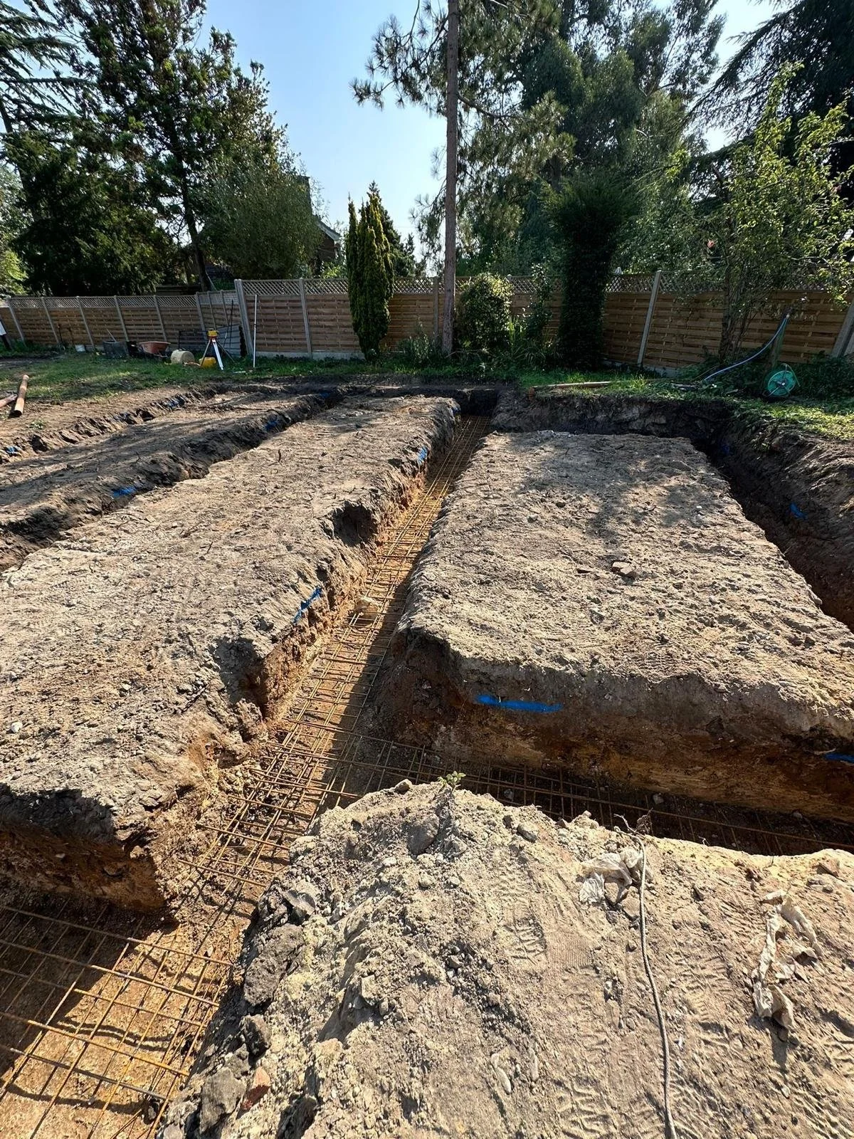 Construction site with dug trenches and exposed rebar for foundation, surrounded by grass, trees, and a wooden fence, under clear blue sky.