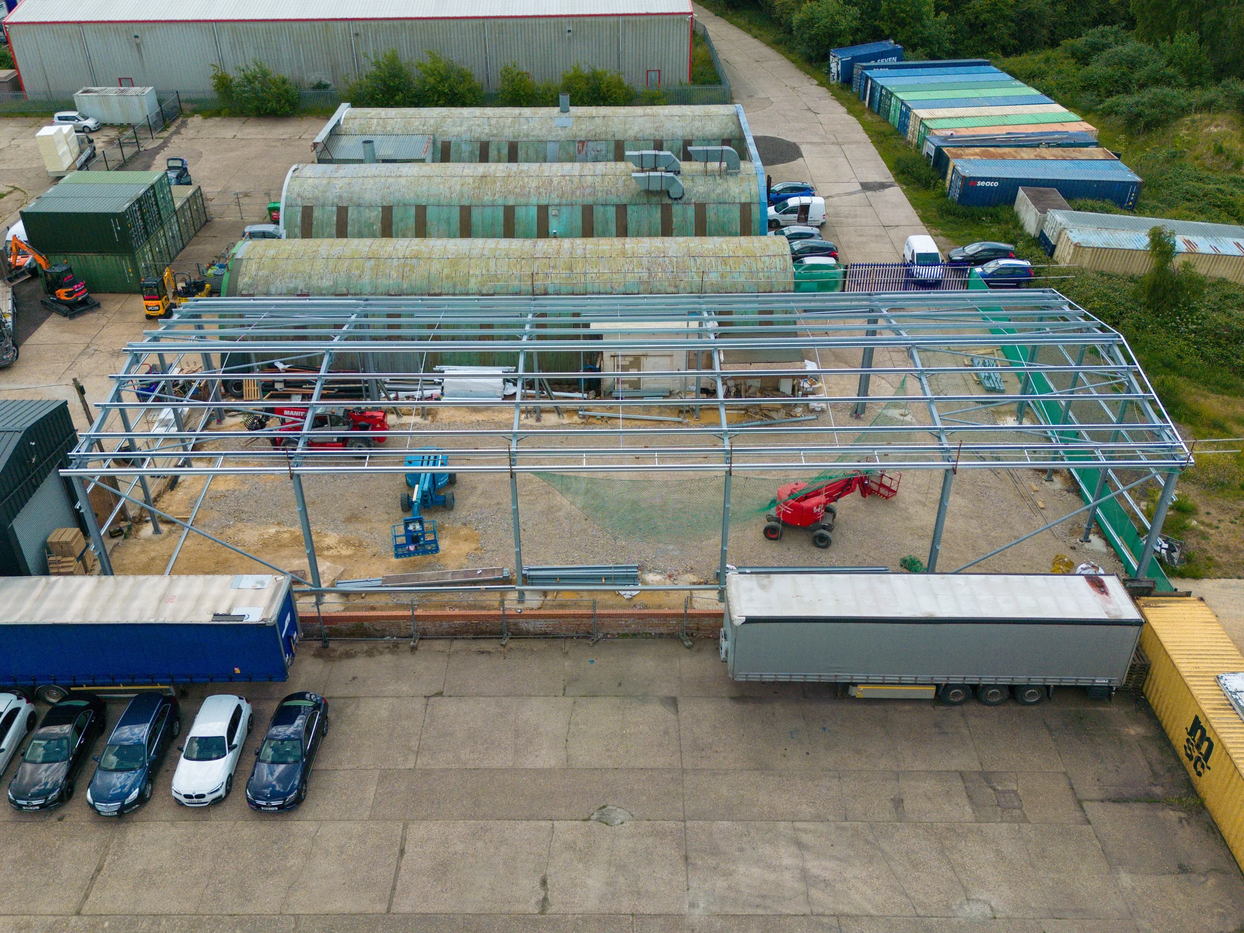 An aerial view of a construction site where a steel framework for a building is being erected. There are construction equipment and vehicles, including a red lift and a blue lift, within the site. Several parked cars and trucks are nearby, along with