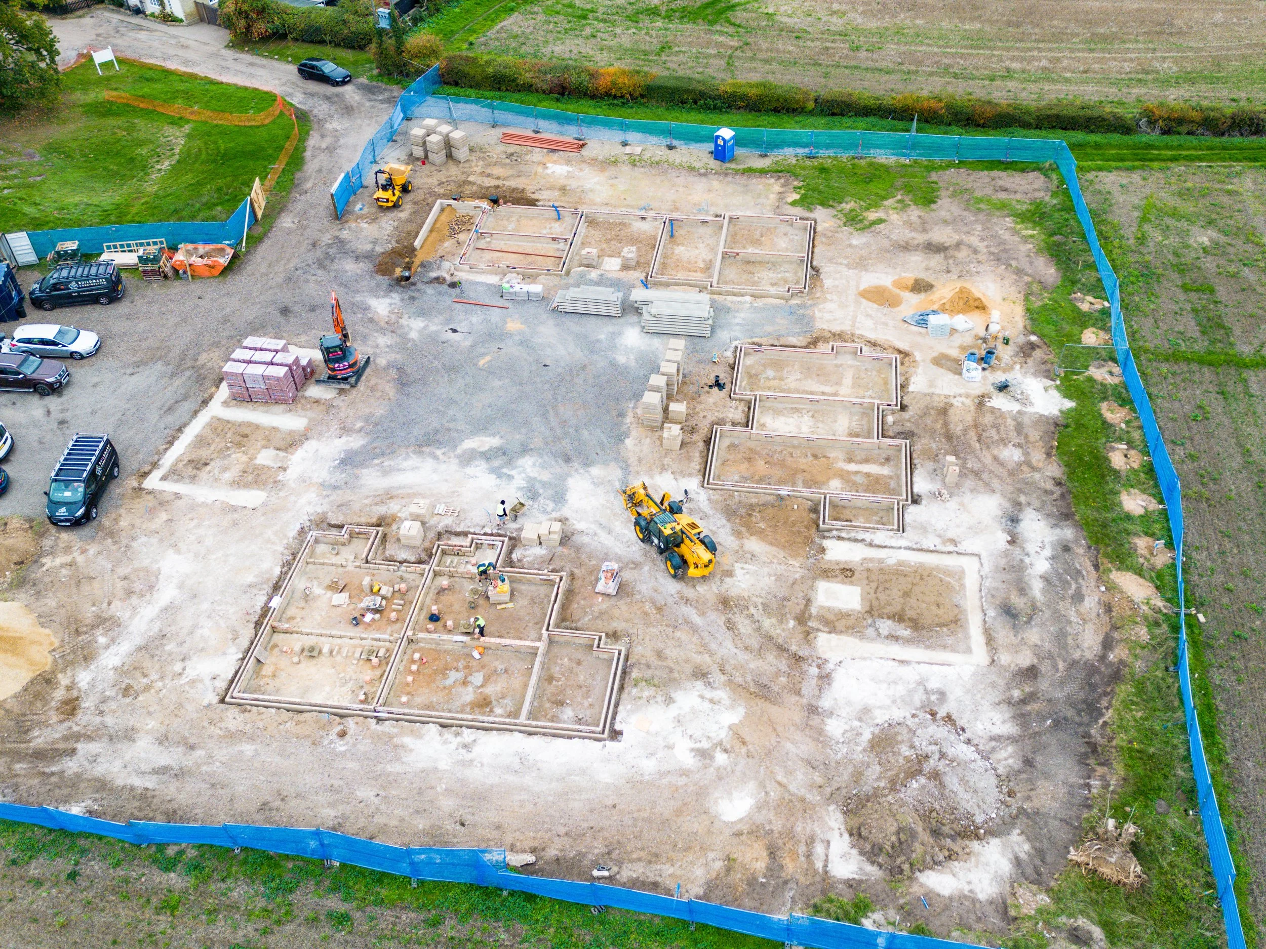 Aerial view of a construction site with foundation outlines, construction equipment, and parked vehicles, enclosed by a blue safety fence.
