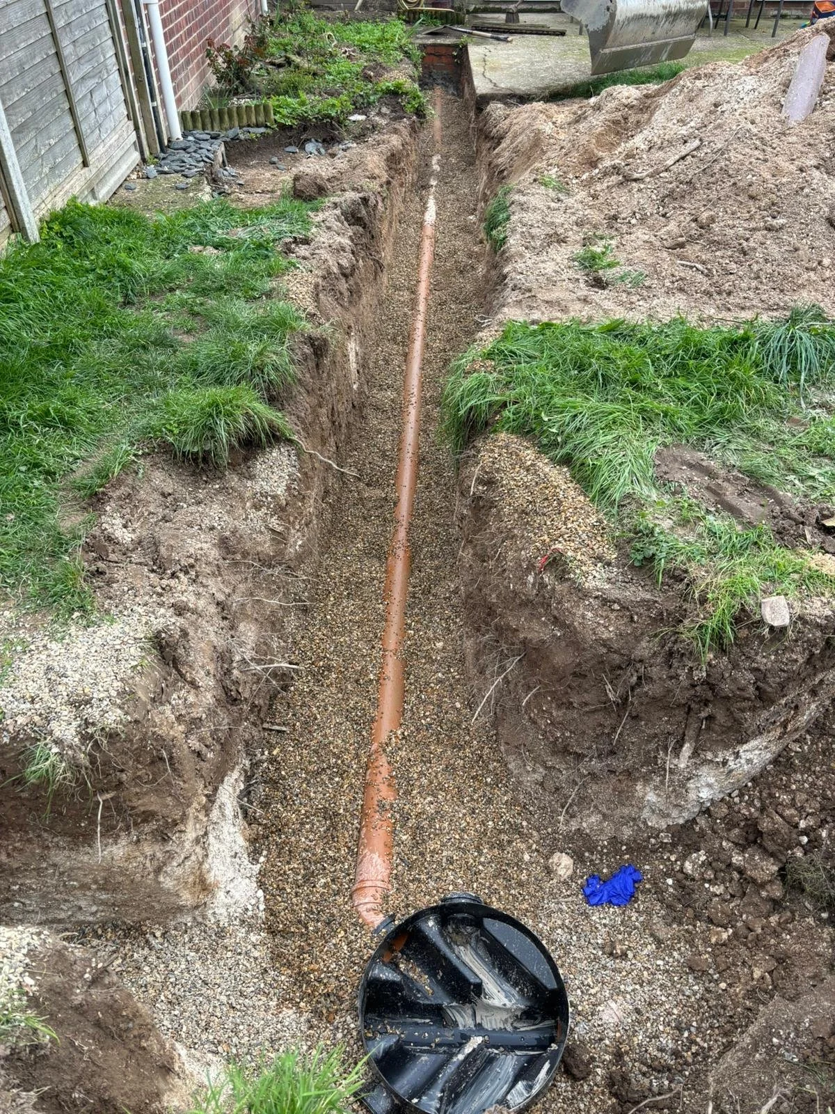 A trench with a pipe being installed in the ground next to a house, with surrounding dirt and grass.