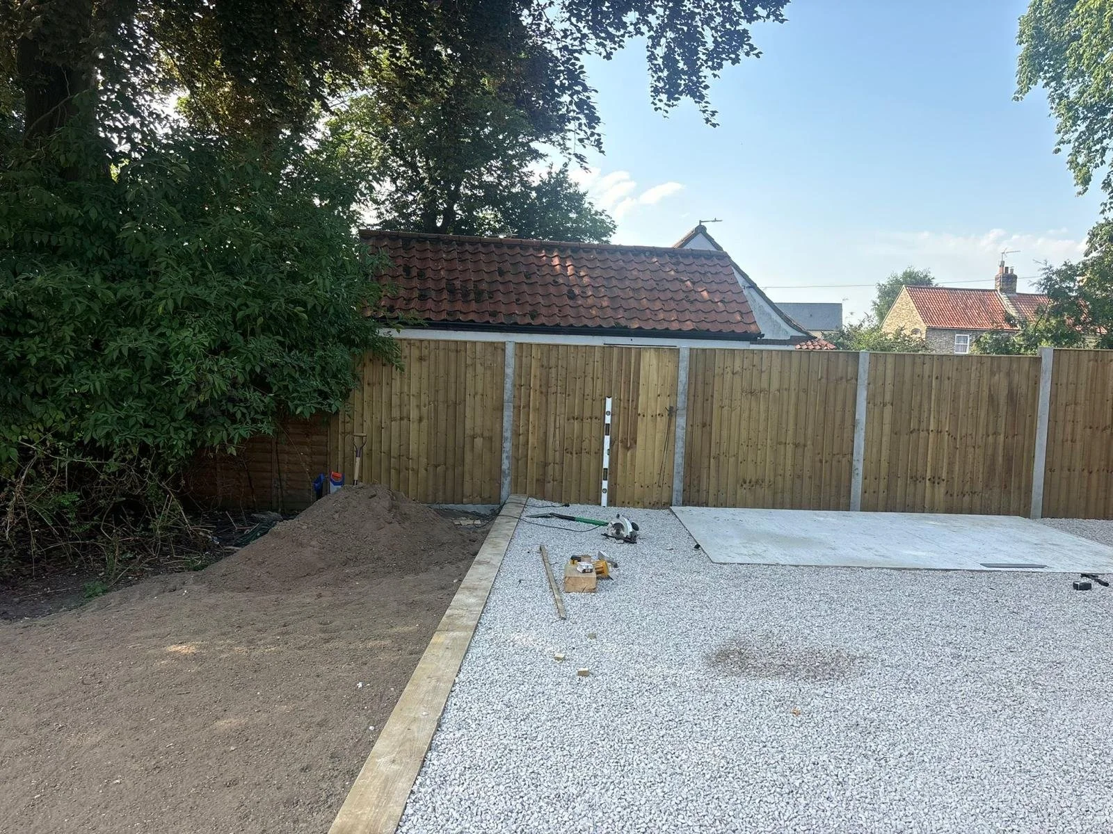 A backyard with a gravel patio area under construction, a wooden fence, and a pile of dirt and construction tools on the ground.
