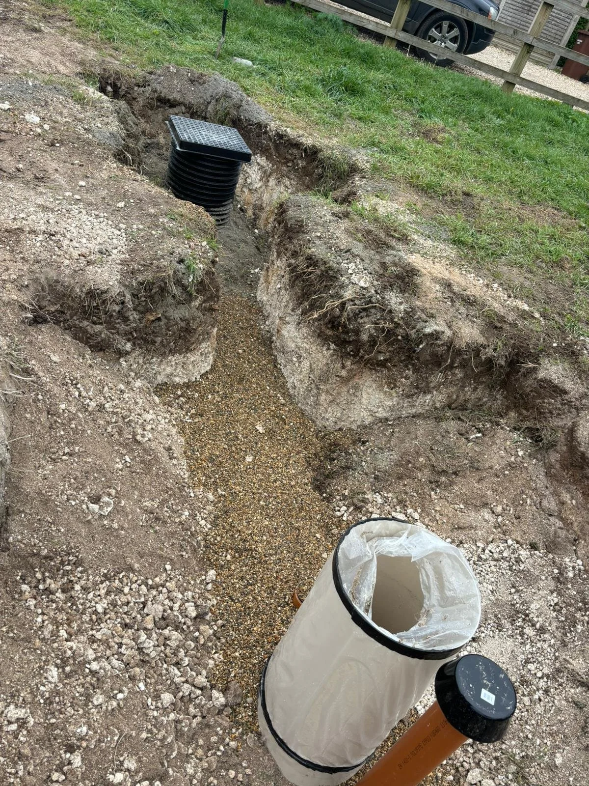 Excavated trench with drainage pipes and gravel at construction site, with grass and parked vehicle in background.