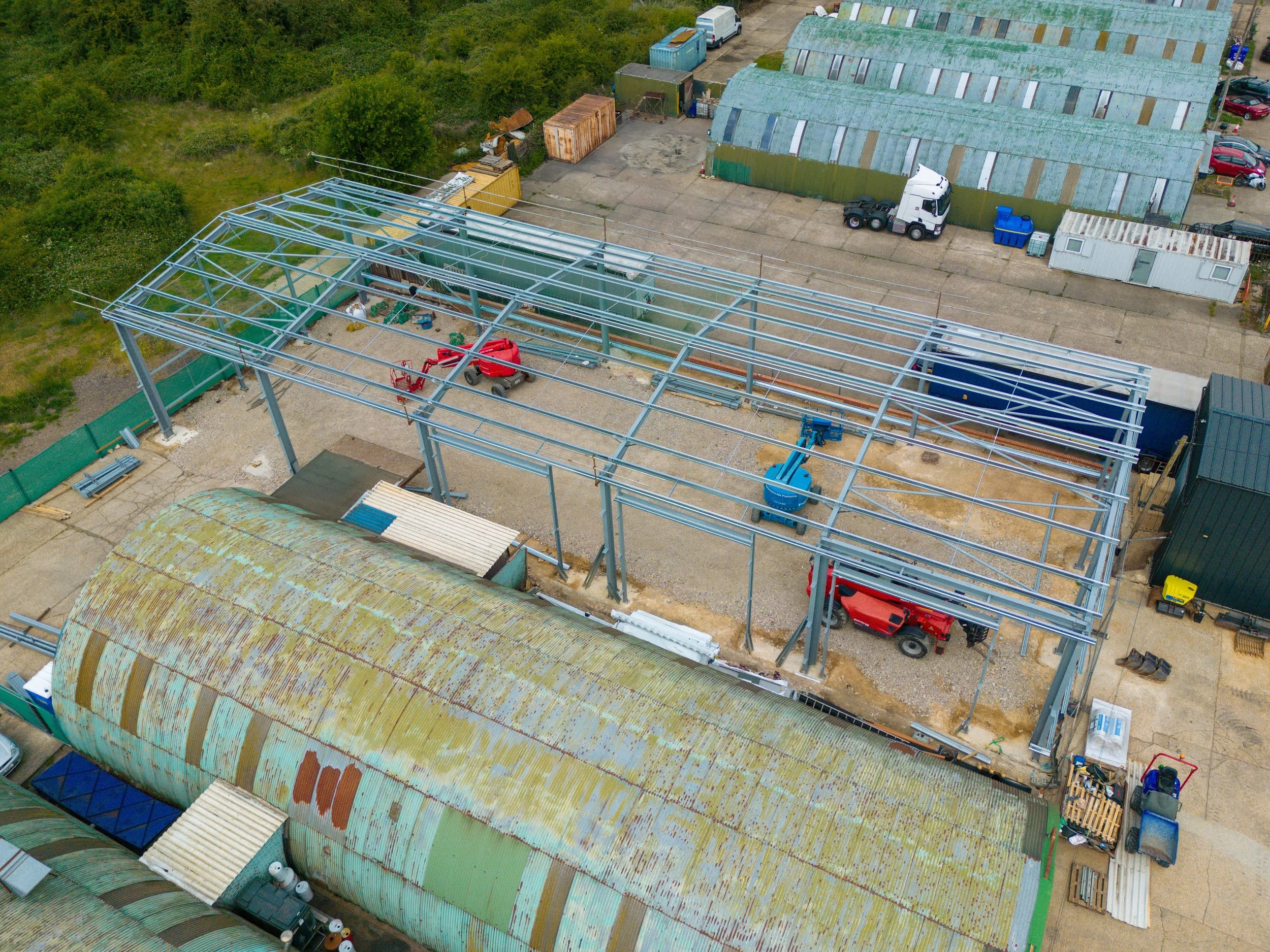 A construction site with a steel framework for a building, construction equipment, and an old rusted green barn roof adjacent to newer green buildings with parked cars, trucks, and containers in the background.