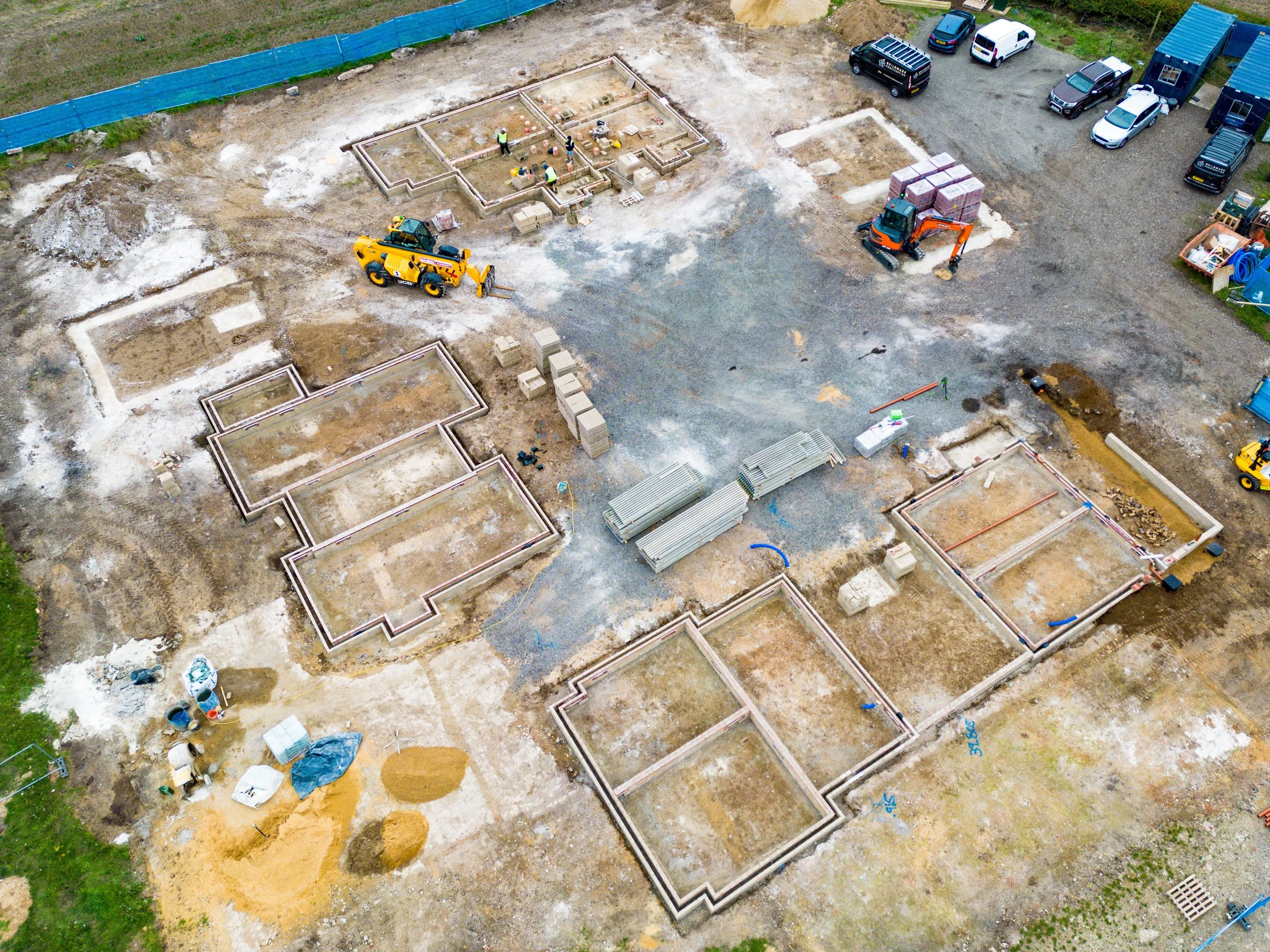 An aerial view of a construction site with foundations being laid, construction workers, machinery, and parked vehicles.