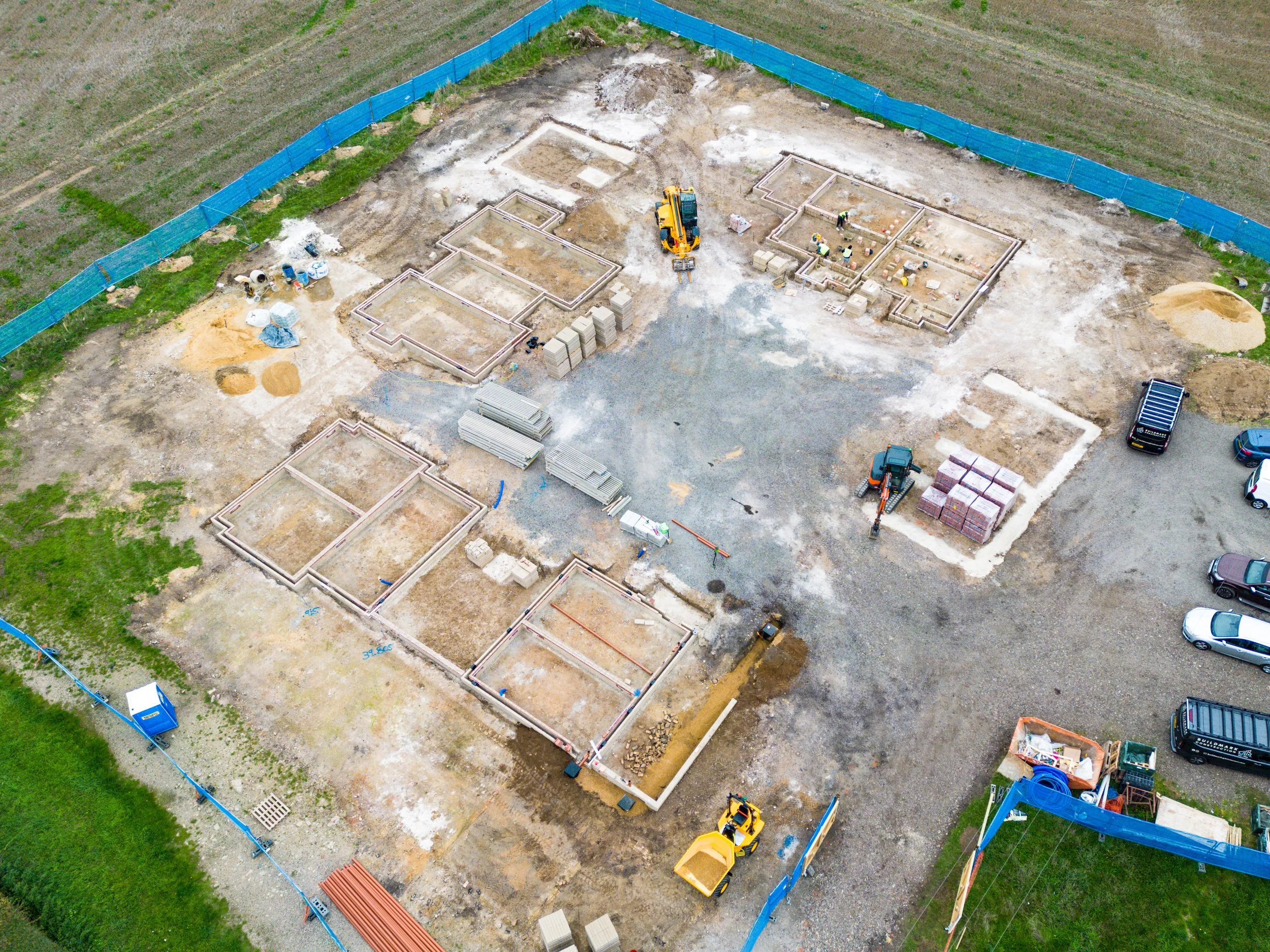 Aerial view of a construction site with partially built foundations, construction equipment, and parked vehicles, surrounded by a blue safety fence.