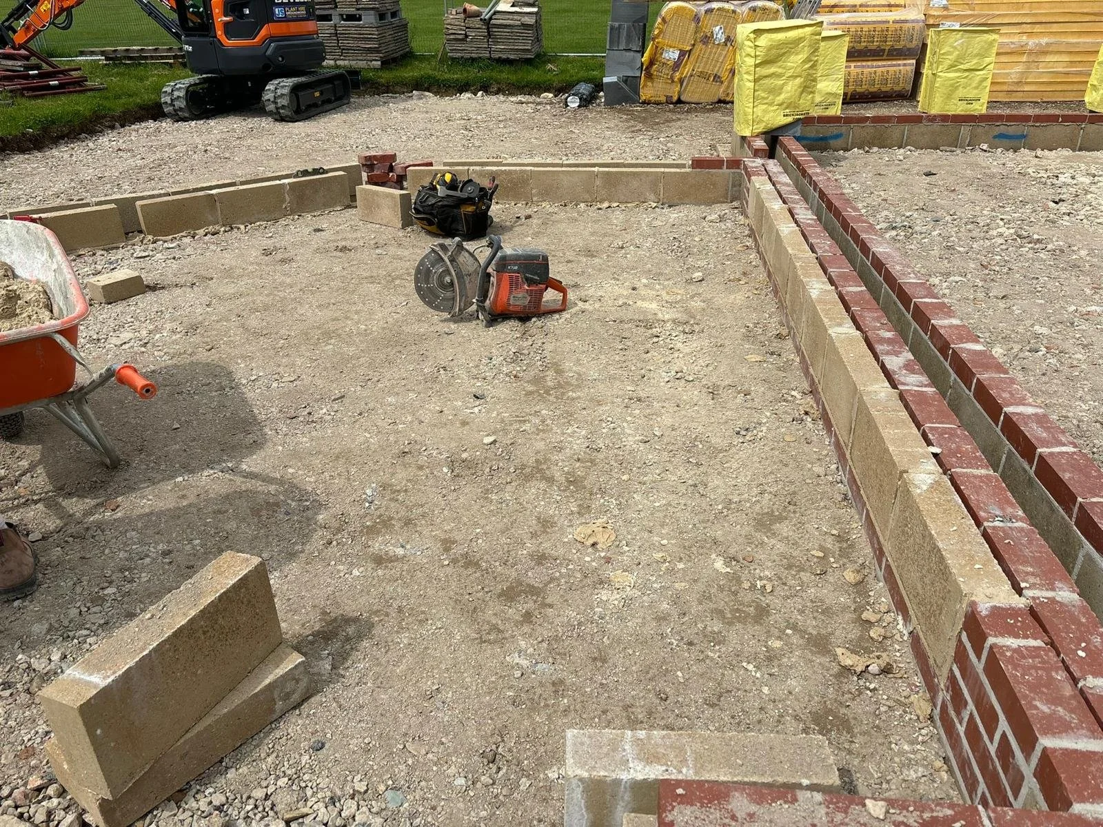 Construction site with brick wall being built, construction tools including a circular saw, a wheelbarrow, and stacks of building materials visible.