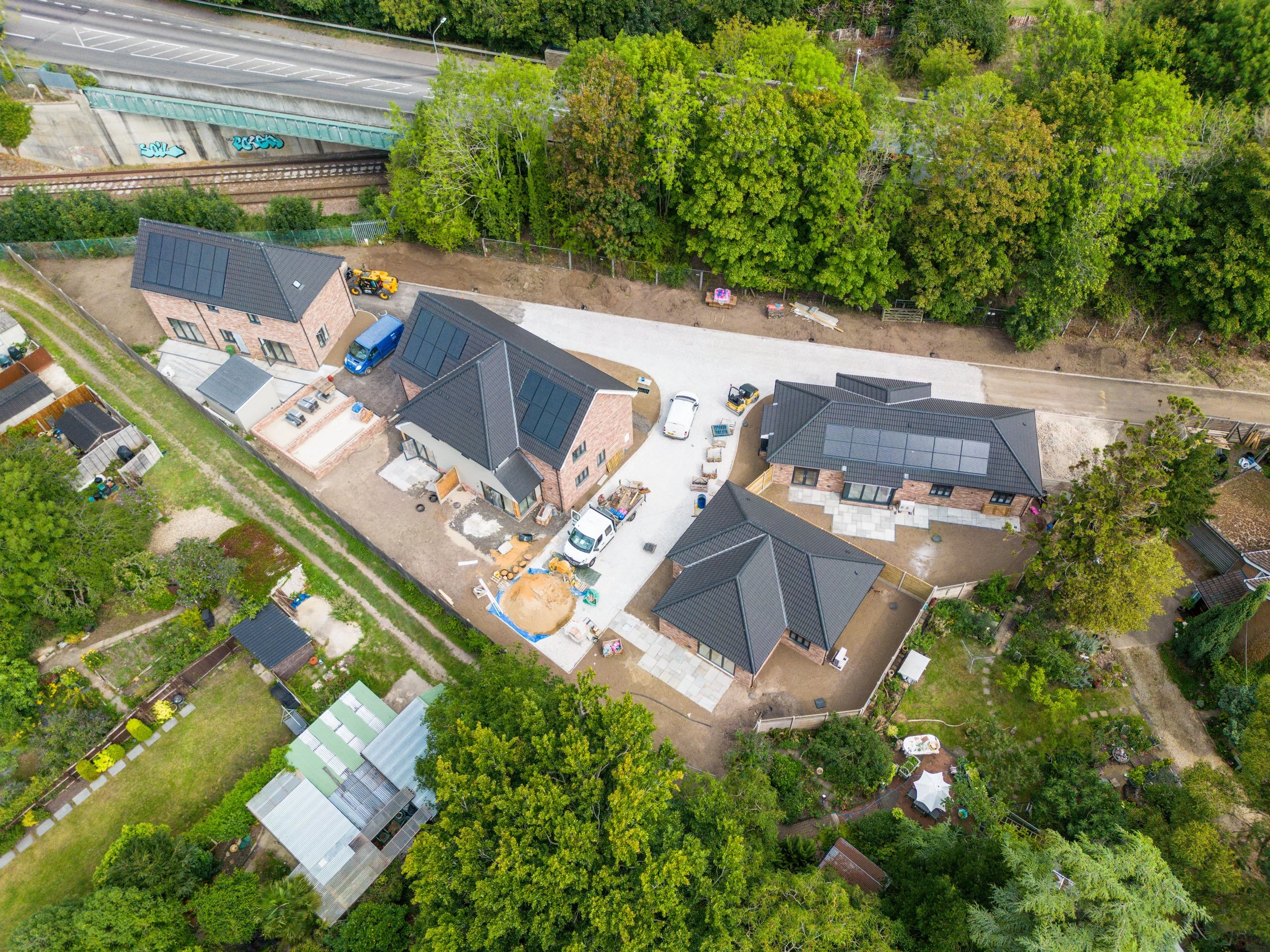 Aerial view of multiple houses under construction with solar panels on rooftops, surrounded by trees and a backyard garden.