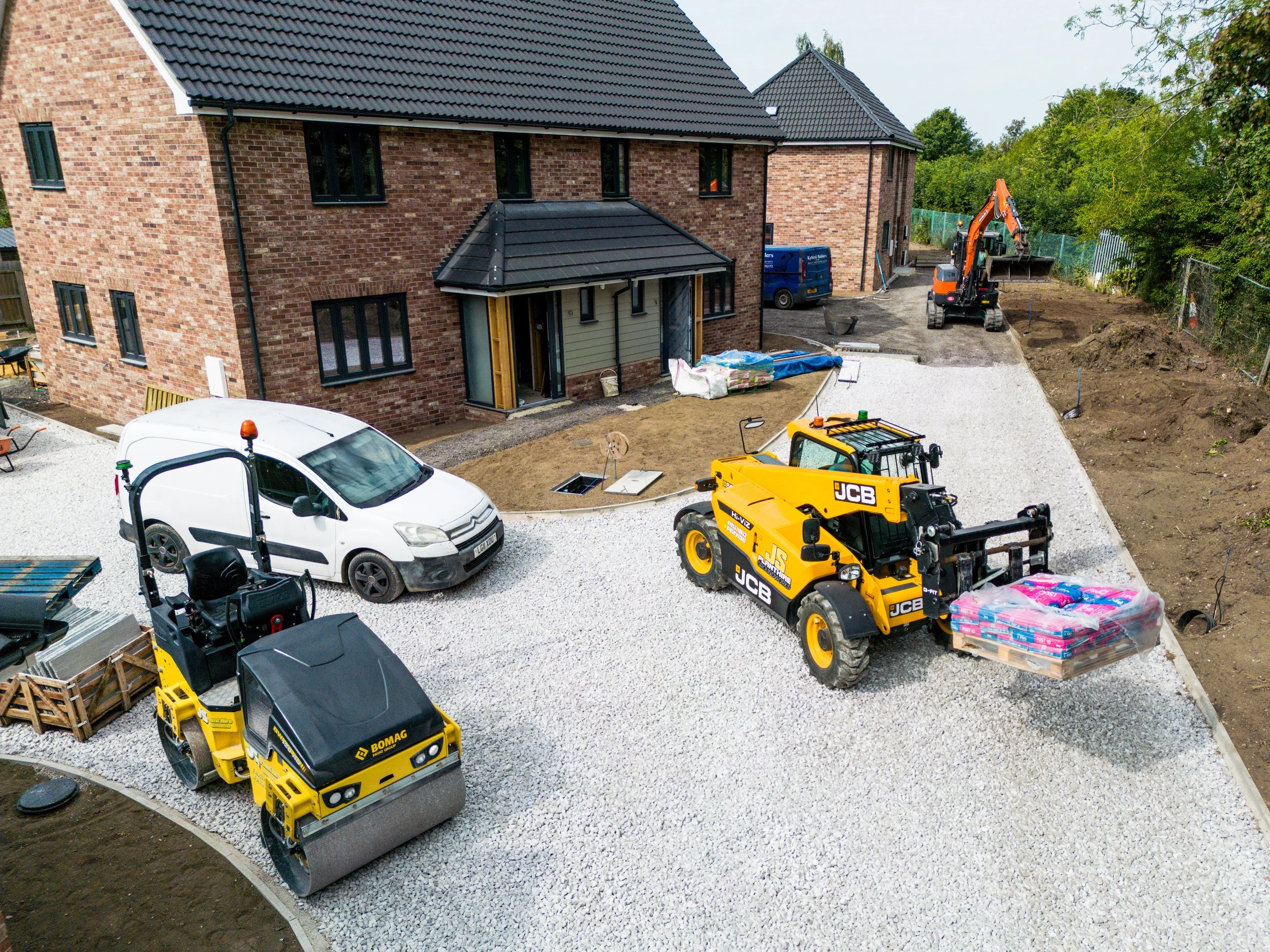 Construction site in front of a brick house with vehicles and machinery, including a road roller, a JCB loader, and a small excavator, with gravel and dirt areas.