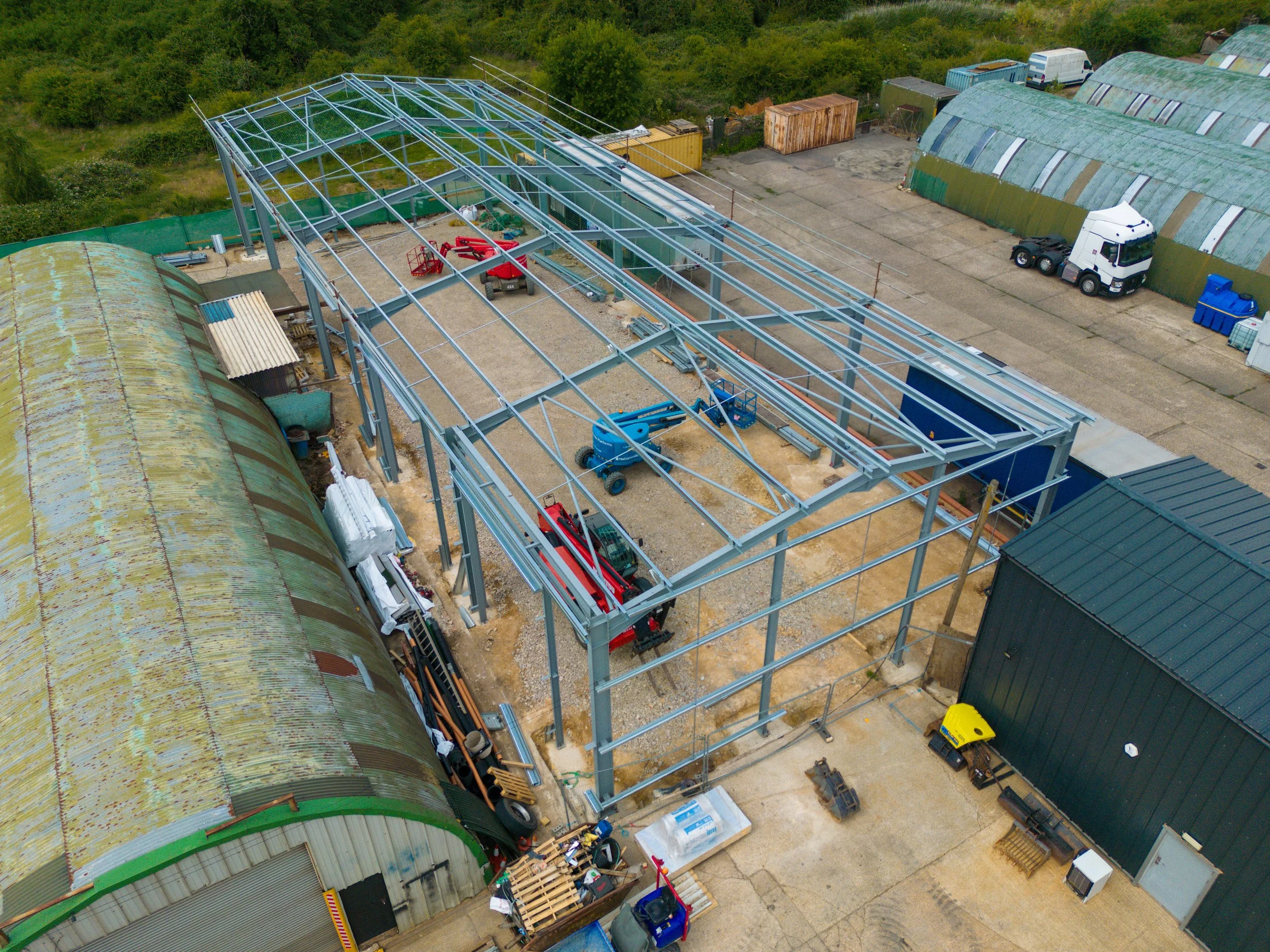 Aerial view of a construction site showing metal framework for a new building, with equipment and materials scattered on the ground, alongside existing greenhouses and a warehouse.