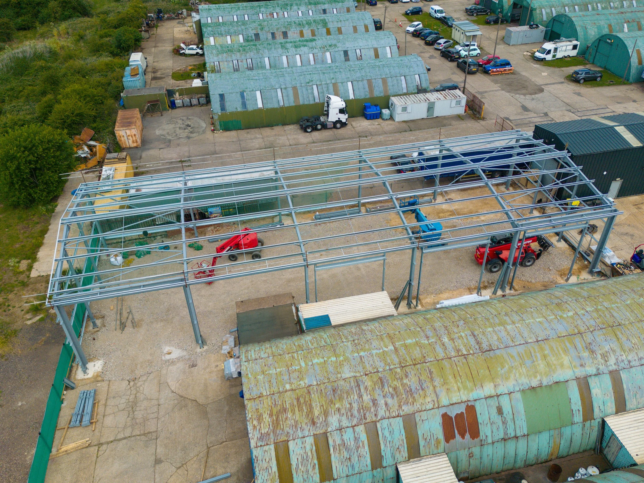 An aerial view of a construction site with a steel frame structure being built inside a partially covered building. Construction vehicles and equipment are present on the site, with construction materials and debris scattered around. There are greenh