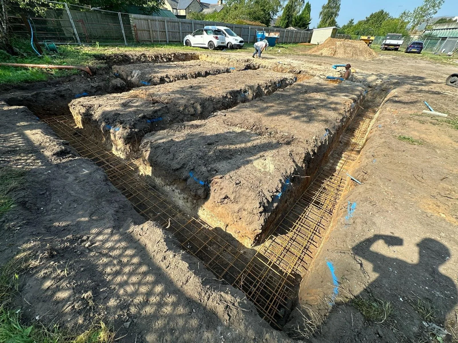 Construction site with excavated trenches and rusted rebar rebar reinforcing the foundation, with workers and parked cars in the background.