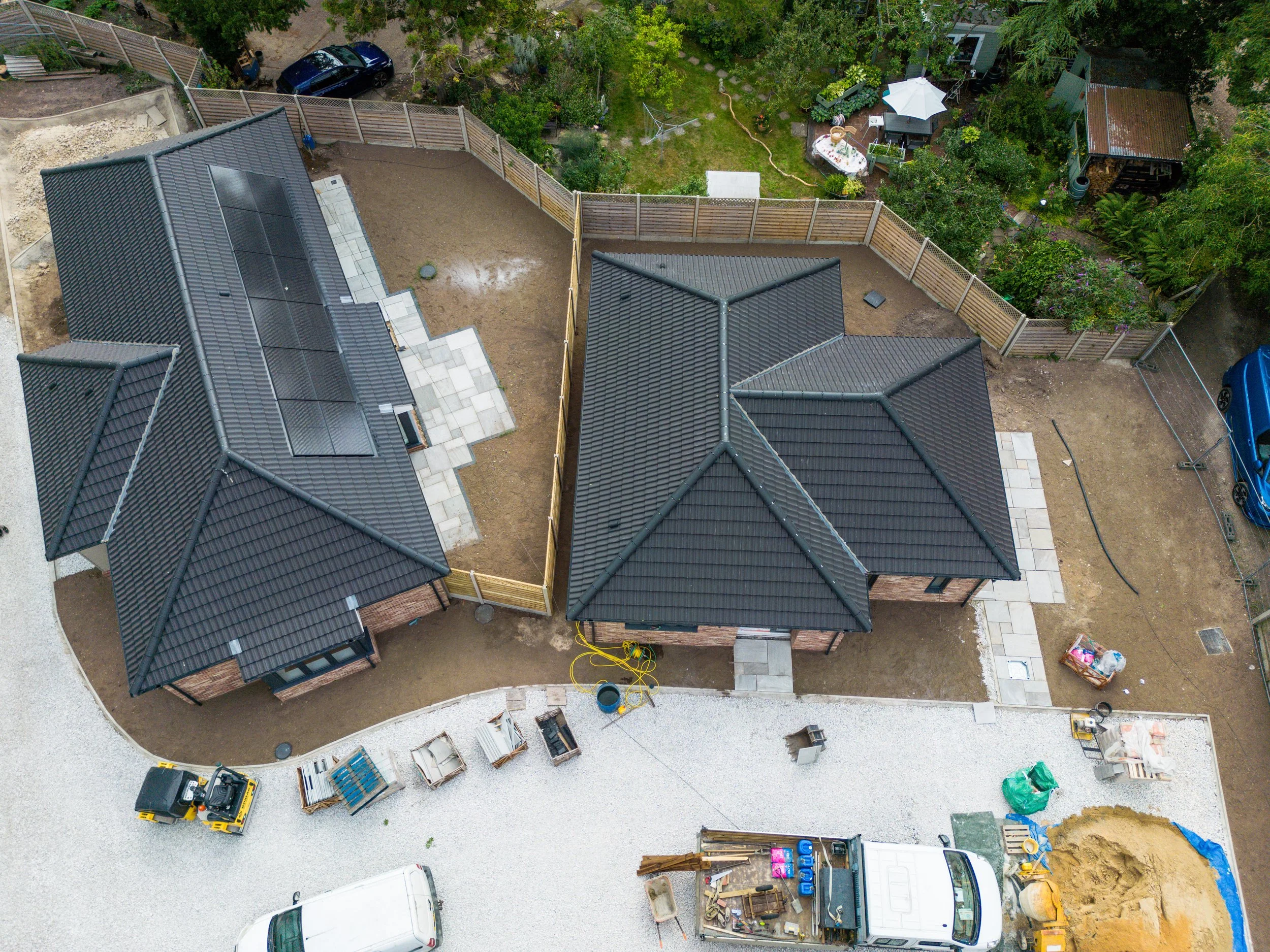 An aerial view of a residential backyard under construction, featuring two houses with black tiled roofs, a dirt yard, a gravel driveway with parked vehicles, construction materials, and a fenced garden area with green trees and outdoor furniture.