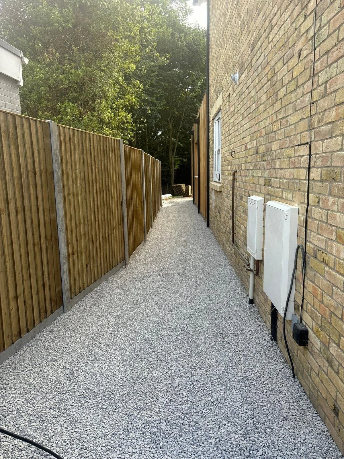 Narrow backyard with gravel pathway, wooden fence on the left, brick wall on the right, and trees in the background.