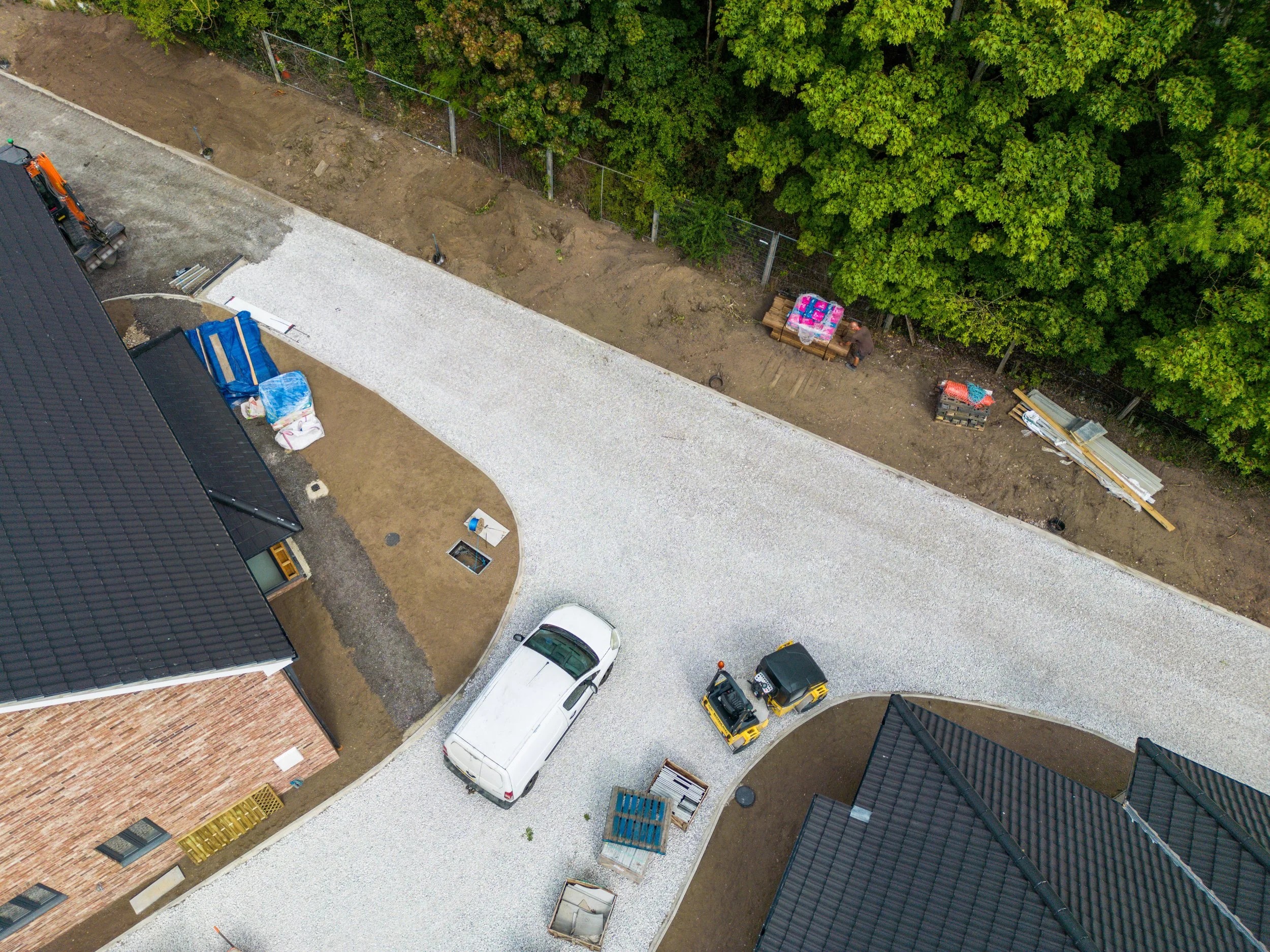 An aerial view of a residential construction site showing a curved gravel driveway, partially built houses with tiled roofs, construction materials, and machinery. There are green trees in the background.