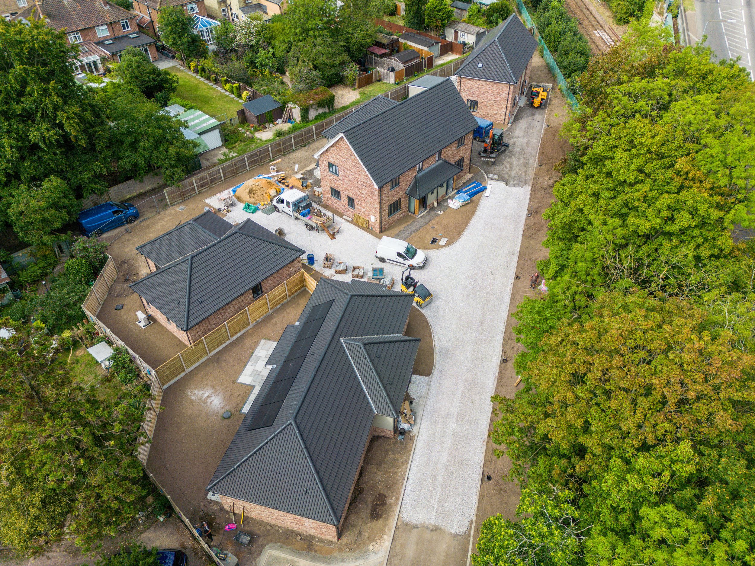 Aerial view of a residential construction site showing houses, vehicles, construction materials, and workers. Completed houses with dark roofs are visible, along with unpaved areas and a newly paved driveway. Surrounding trees and neighboring homes a