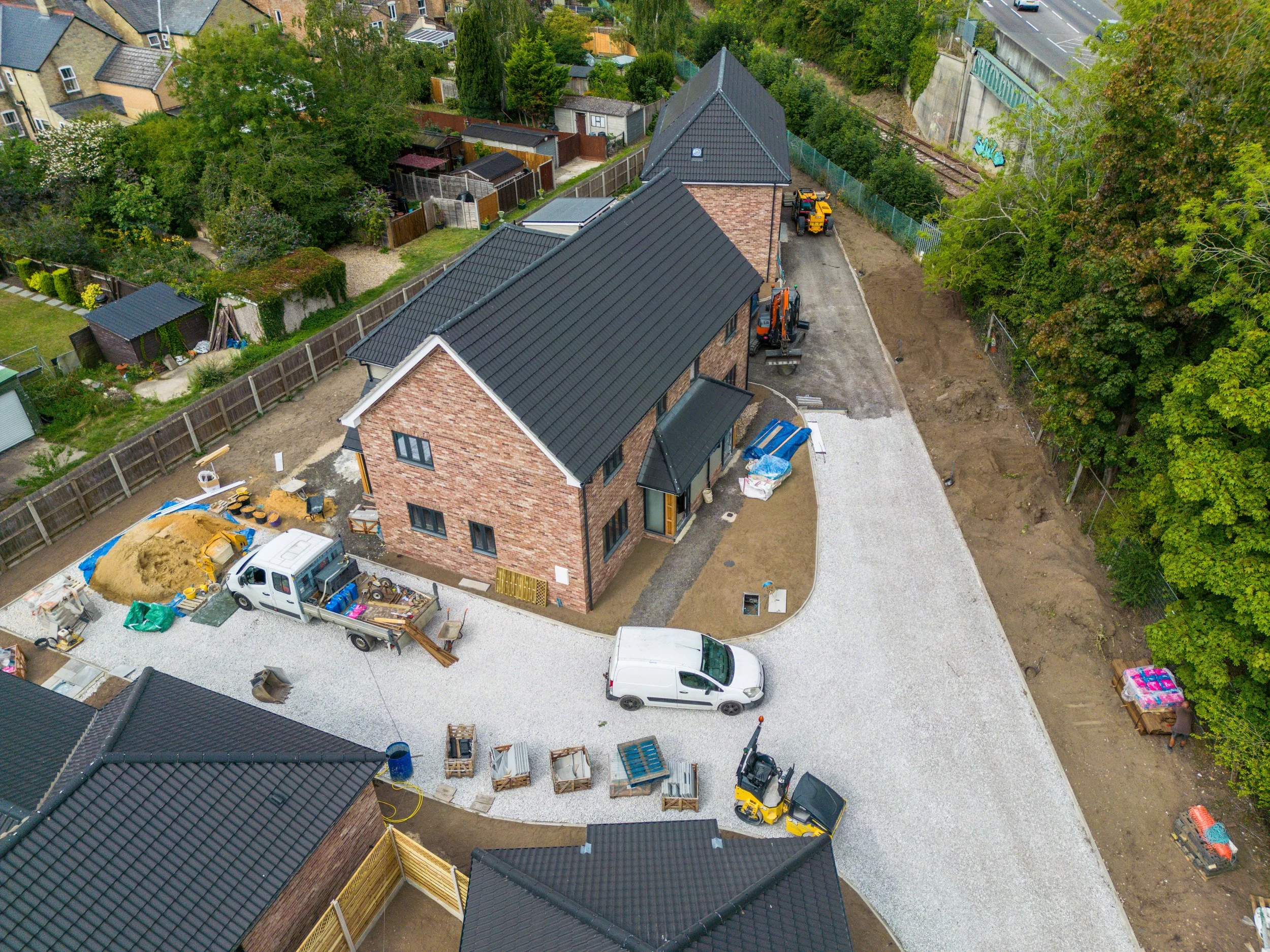 Aerial view of a house under construction with gravel driveway, construction vehicles, and building materials. Neighbors' houses and greenery are visible around.