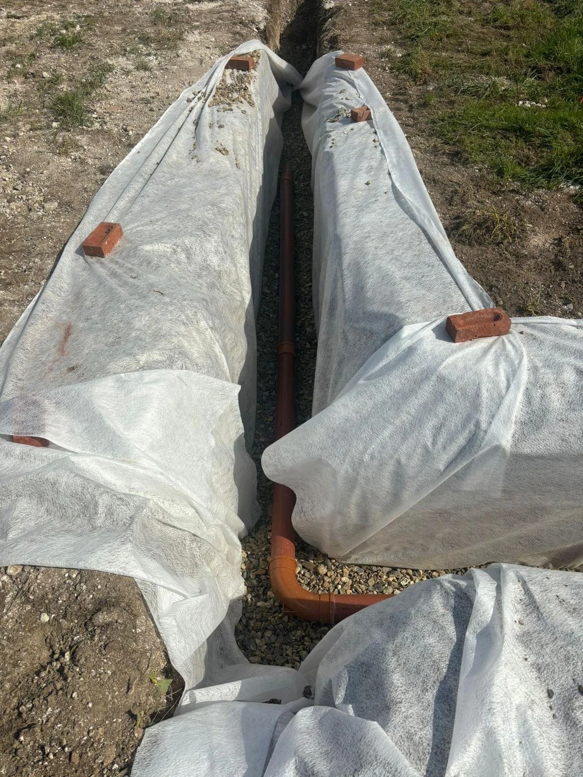 View of two underground pipes covered with white fabric, secured with bricks, and a brown pipe running between them on a patch of dirt and gravel.