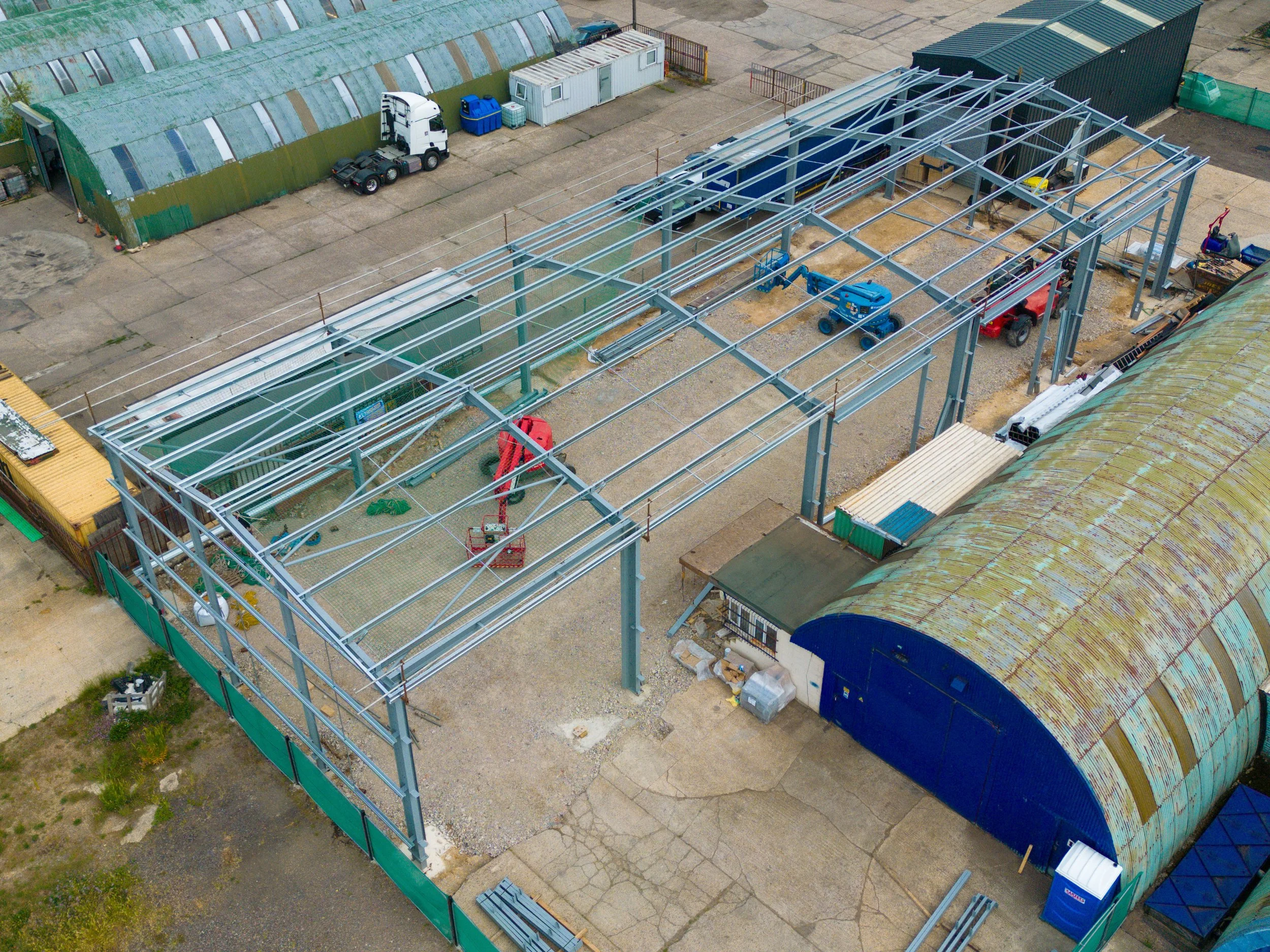 Aerial view of a construction site with steel framework for a building, construction equipment, gravel ground, neighboring green and rusty metal buildings, and construction materials.