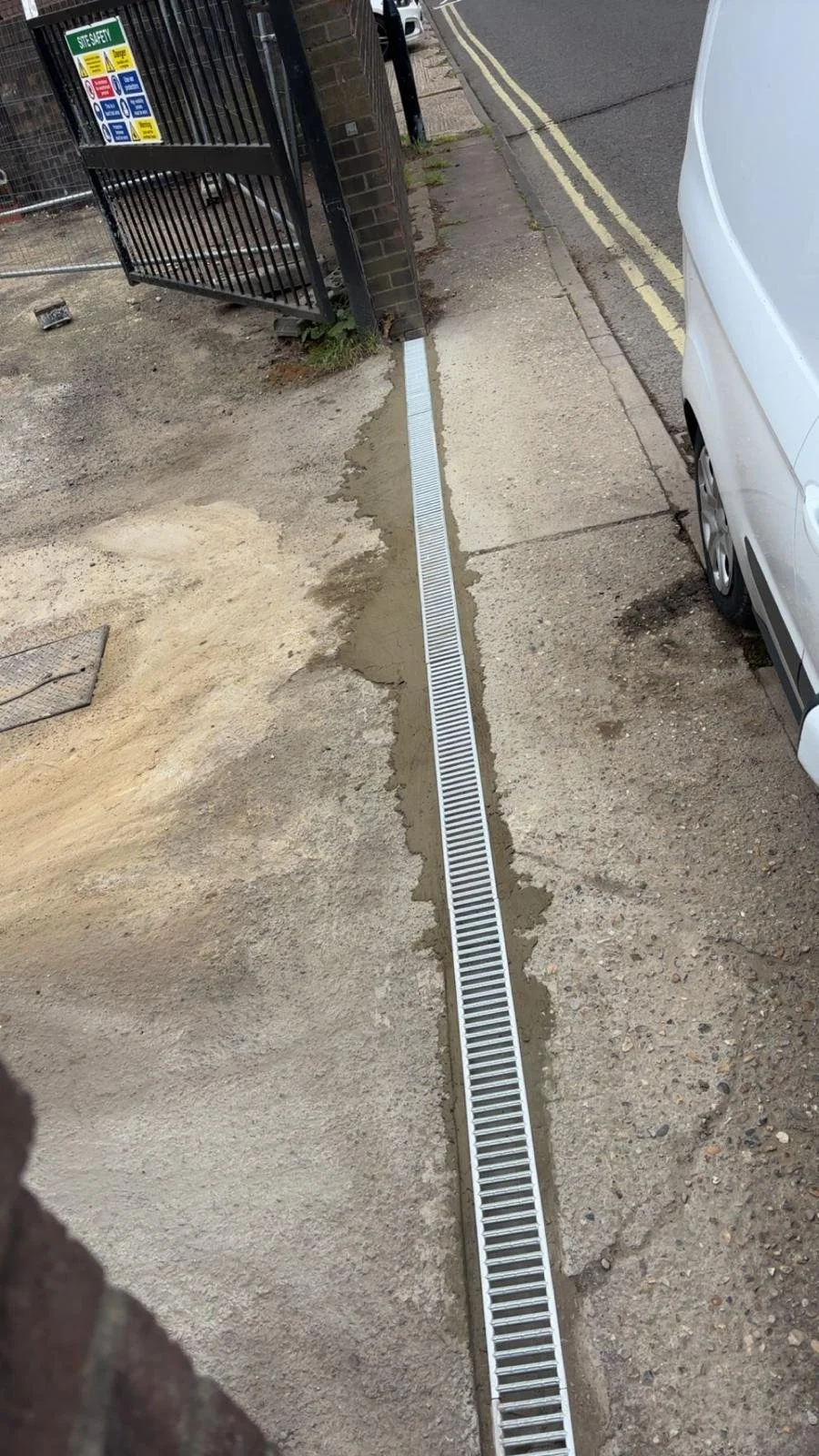 A drainage grate installed along the edge of a sidewalk next to a parked white van and a black gate with a site safety sign, on a street with double yellow lines.