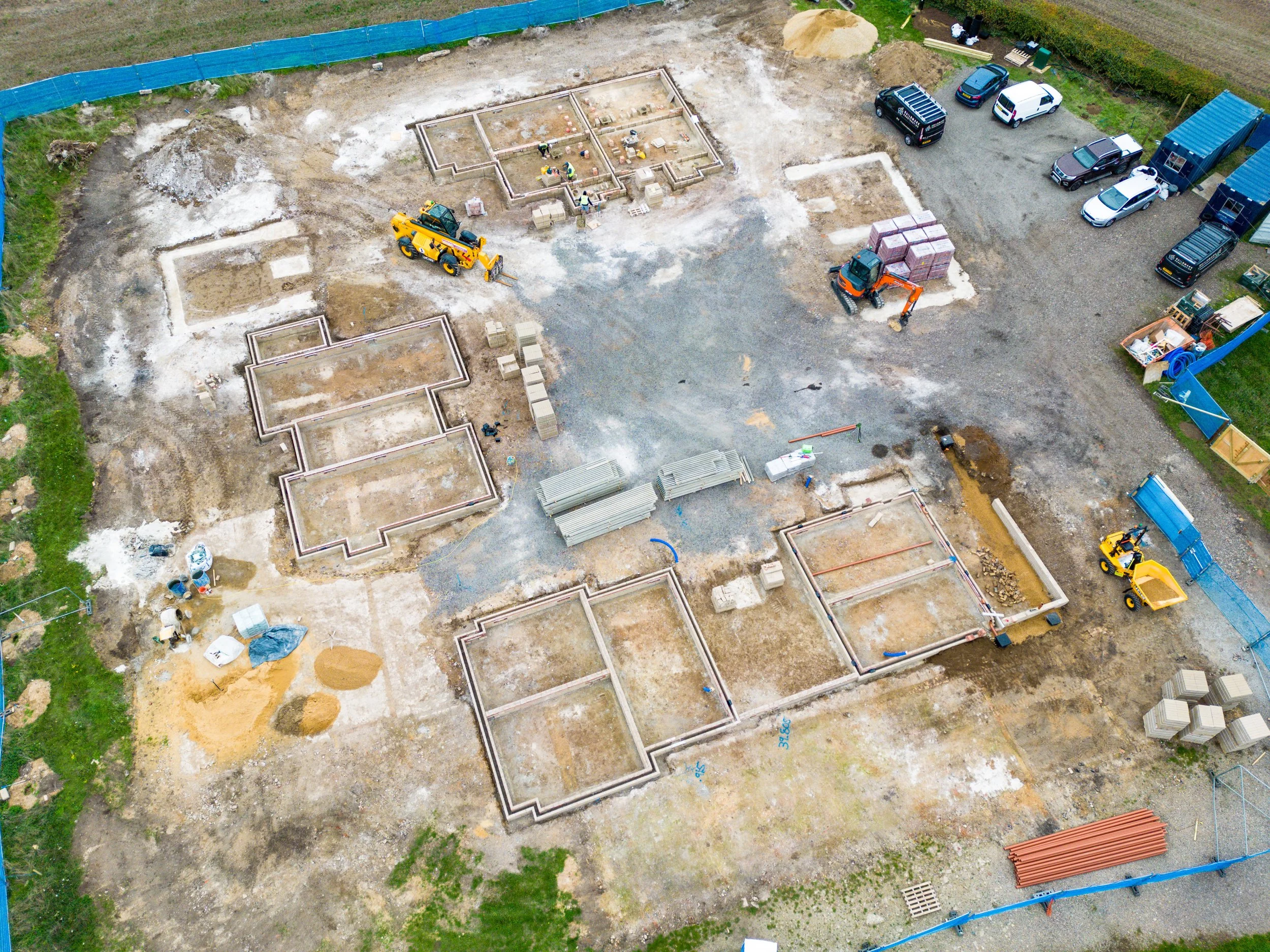 An aerial view of a construction site with partially built foundation walls, construction vehicles, construction materials, and parked cars.