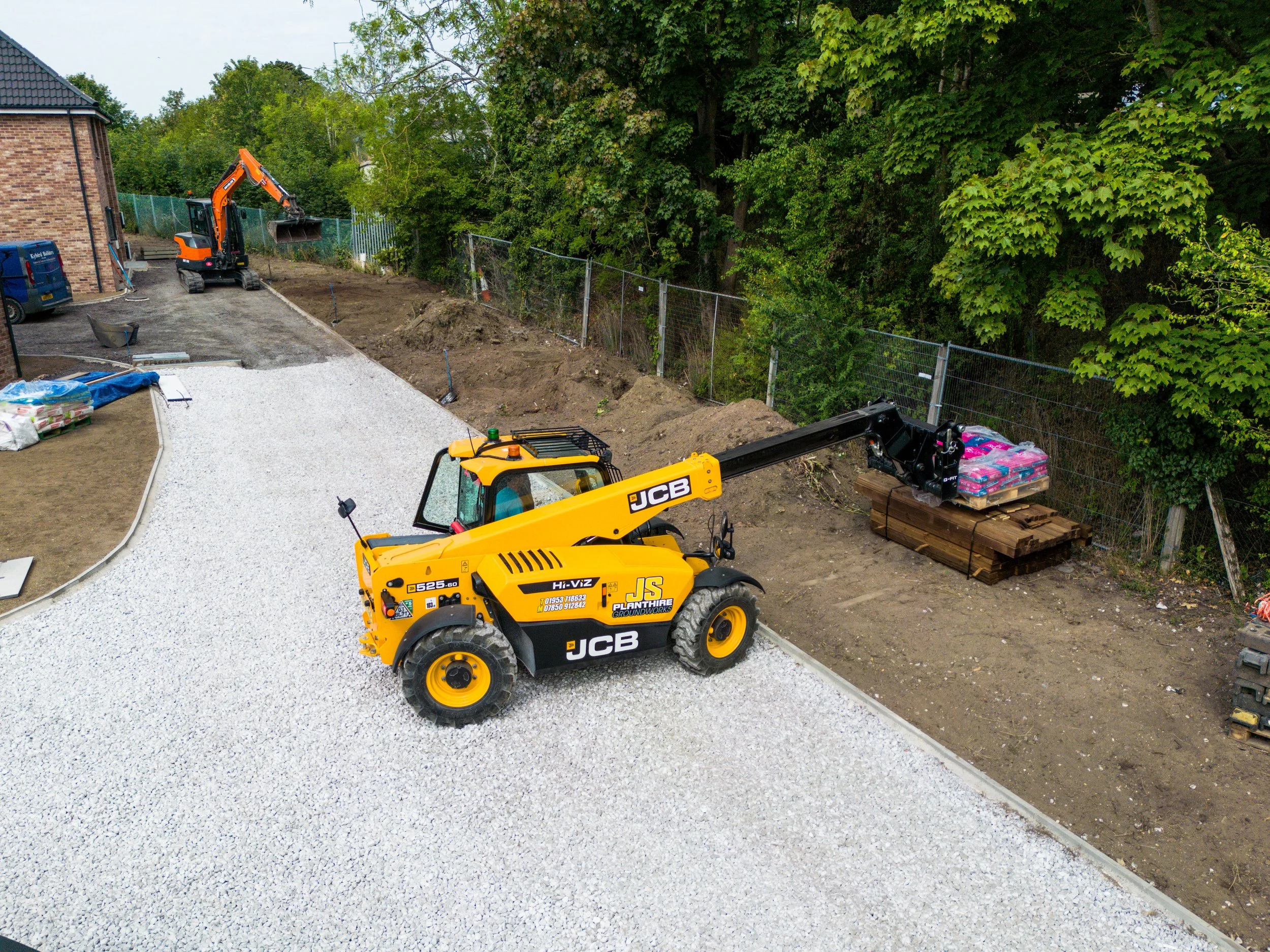 Construction site with a yellow JCB telescopic loader lifting pallets, gravel being laid, and an excavator working in the background near green trees and a fence.