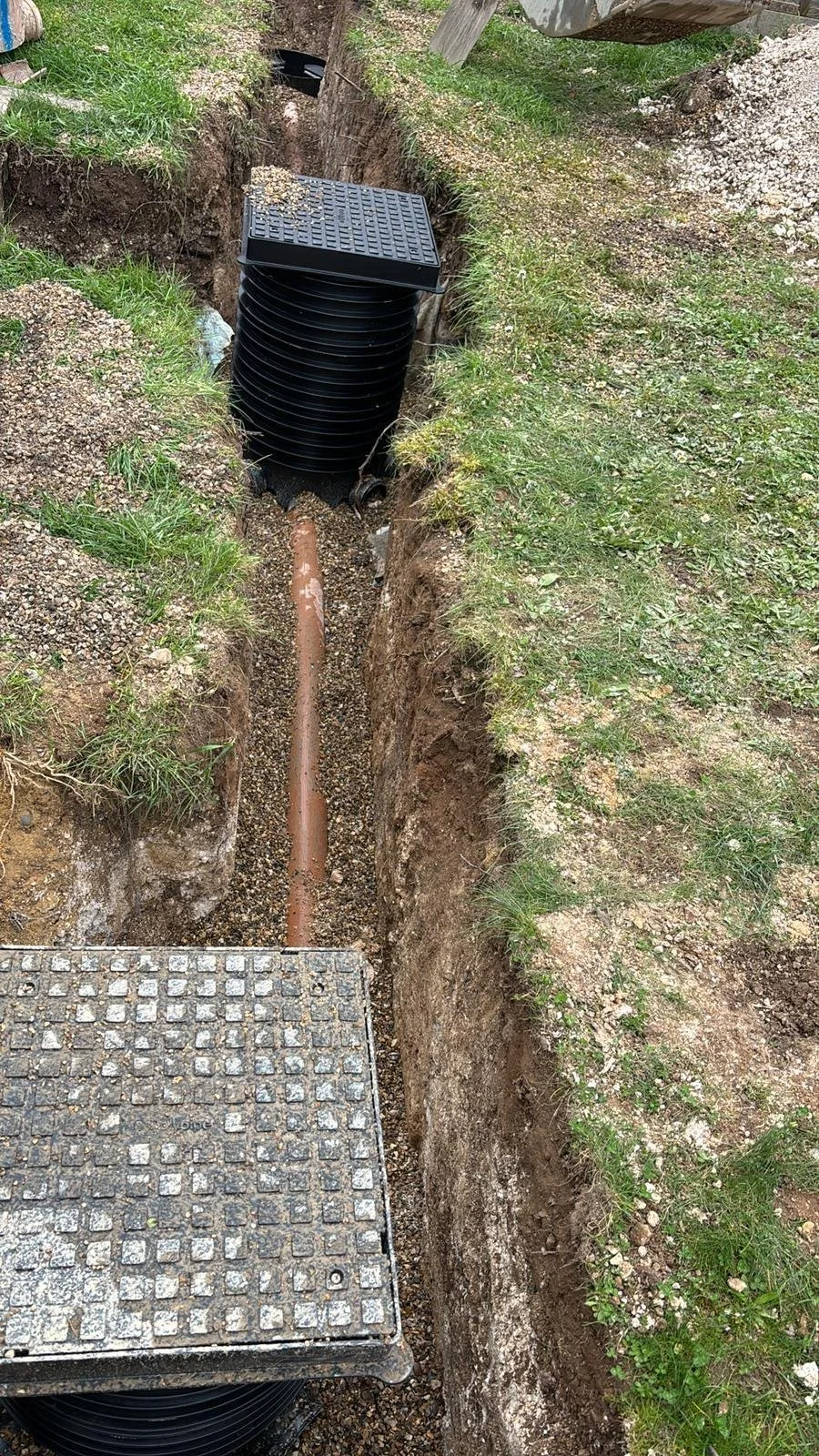 Installation of a underground drainage system with black corrugated pipes, a gravel bed, and a access cover, surrounded by grass and soil.