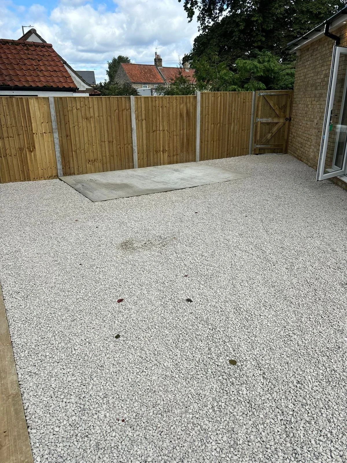 Backyard patio with white gravel surface, enclosed by wooden fence with a small gate, and a brick house wall on the right with a sliding glass door open. Overcast sky with cloudy weather.