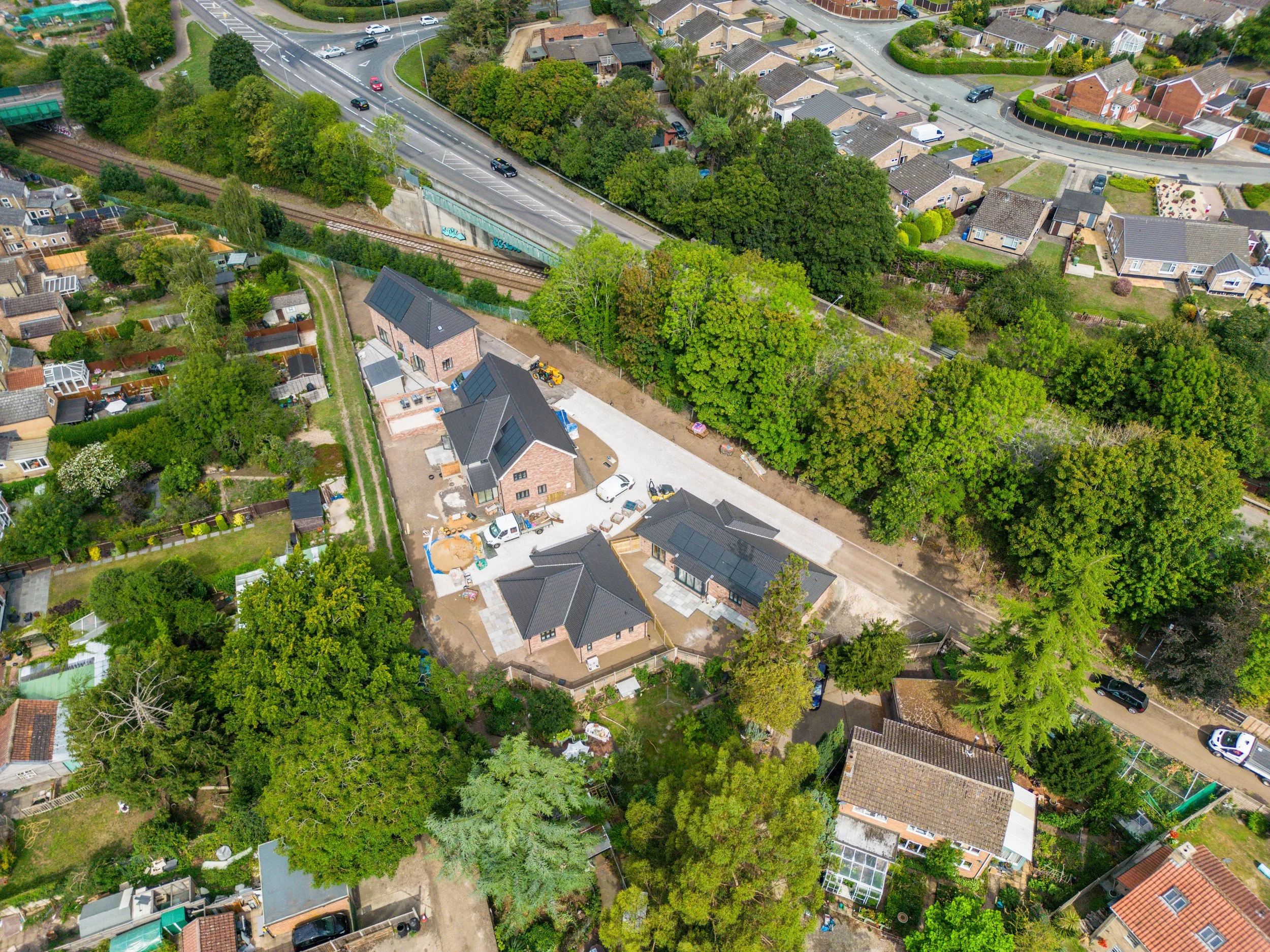 An aerial view of a residential neighborhood with houses, trees, and a railway track. There is a stretch of road with cars and a bridge crossing over the railway. Some houses have solar panels on their roofs, and there are various construction materi