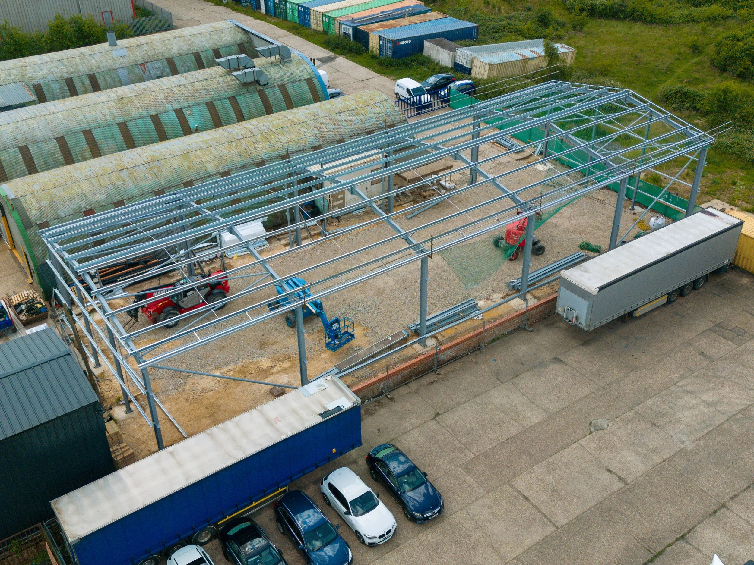 Aerial view of a construction site with a building frame being assembled, surrounded by parked cars and nearby greenhouses and containers.