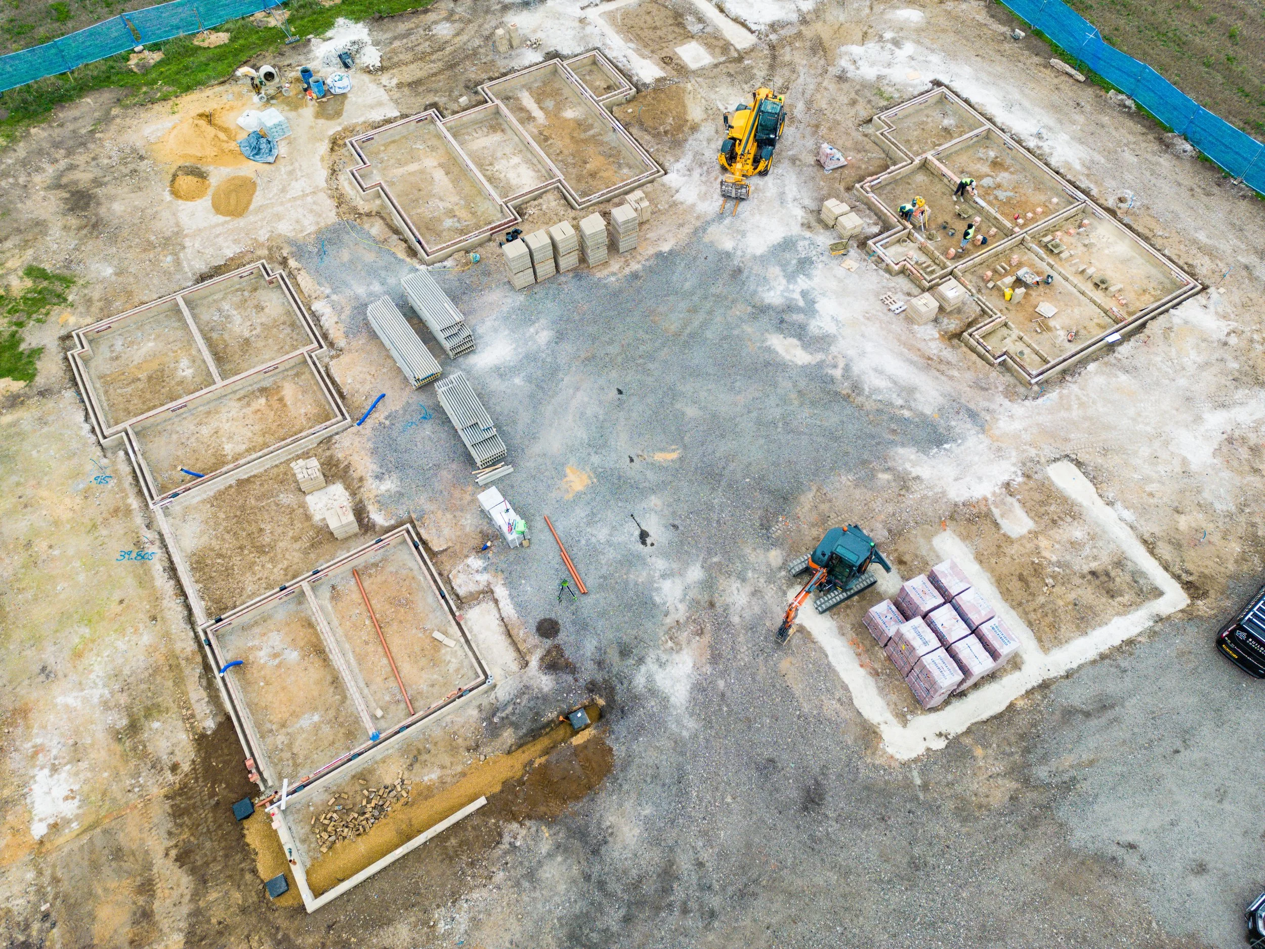 An aerial view of a construction site with six foundation outlines, construction materials, workers, and machinery, including small excavators, within a fenced perimeter.